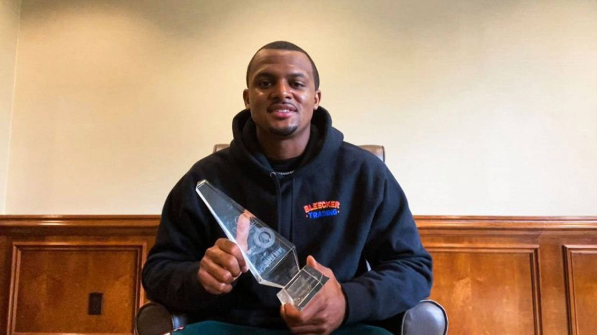 Deshaun Watson 桌布 壁紙 — 一個微笑的男人穿著黑色連帽衫，坐在木質鑲板的房間裡，手持一個透明玻璃獎項。