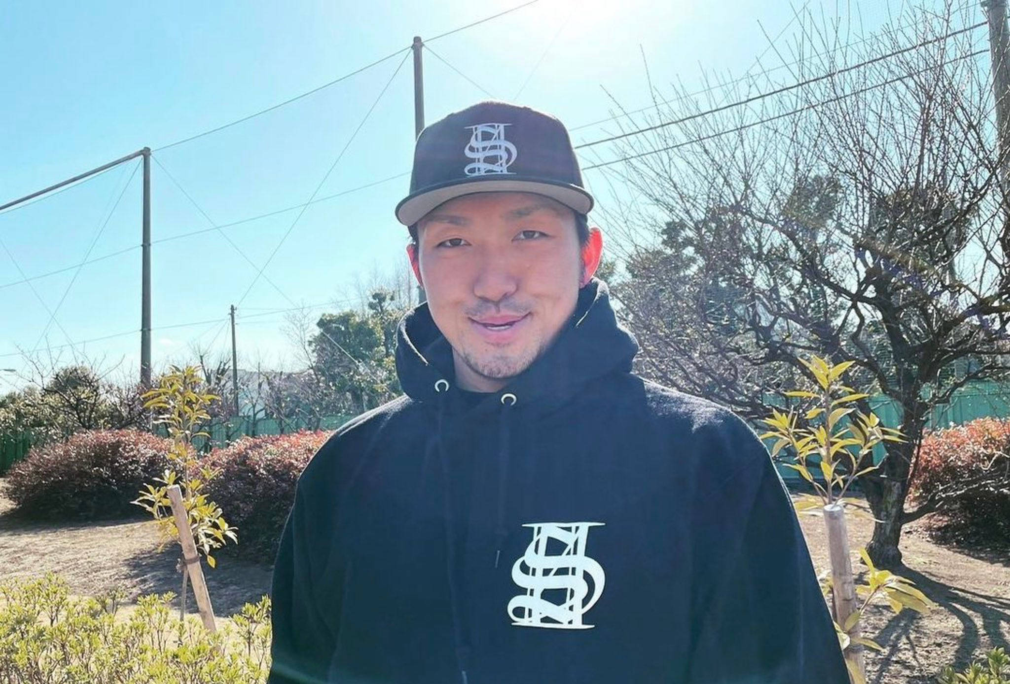 Athlete 桌布 壁紙 — 一個微笑的男人穿著黑色連帽衫和帽子，站在戶外明亮的陽光下，身後是光禿禿的樹木和灌木。