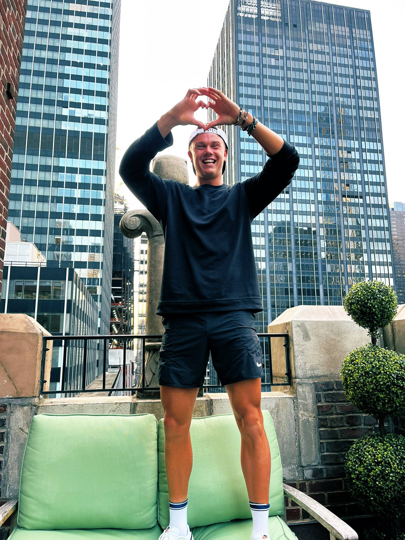 Tennis 桌布 壁紙 — 一個微笑的男人站在天台沙發上，雙手比心，背景是高聳的玻璃摩天大樓。