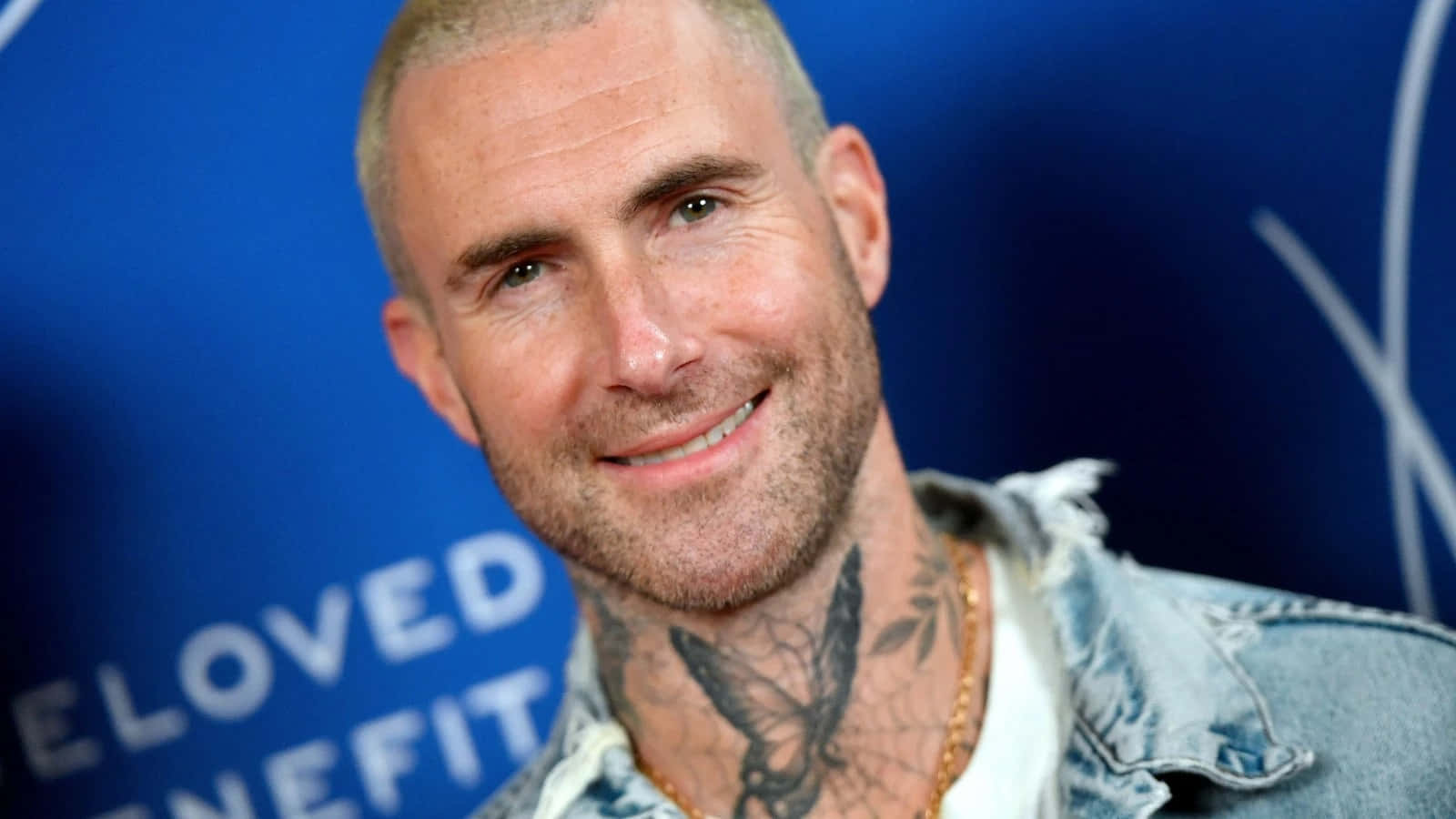 Adam Levine 桌布 壁紙 — 一個微笑的男人近距離肖像，頸上有刺青，穿著牛仔外套，背景為藍色。