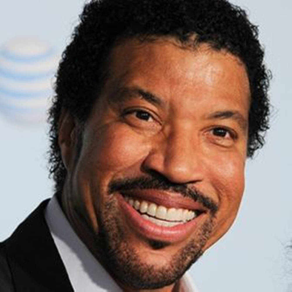 Celebrity 桌布 壁紙 — 一個微笑的男士近距離肖像，他有捲髮和鬍子，穿著西裝，背景是淺藍色。