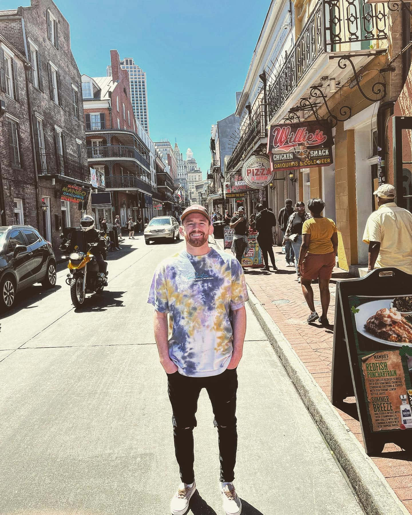 New Orleans 桌布 壁紙 — 一個微笑的男子穿著紮染襯衫，站在一條陽光明媚、熙熙攘攘的街道上，兩旁是歷史建築和美食車。