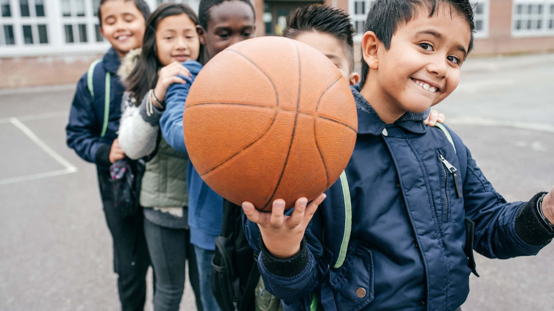 Best Sports 桌布 壁紙 — 一個微笑的男孩拿著籃球，朋友們聚集在他身後的學校球場上。