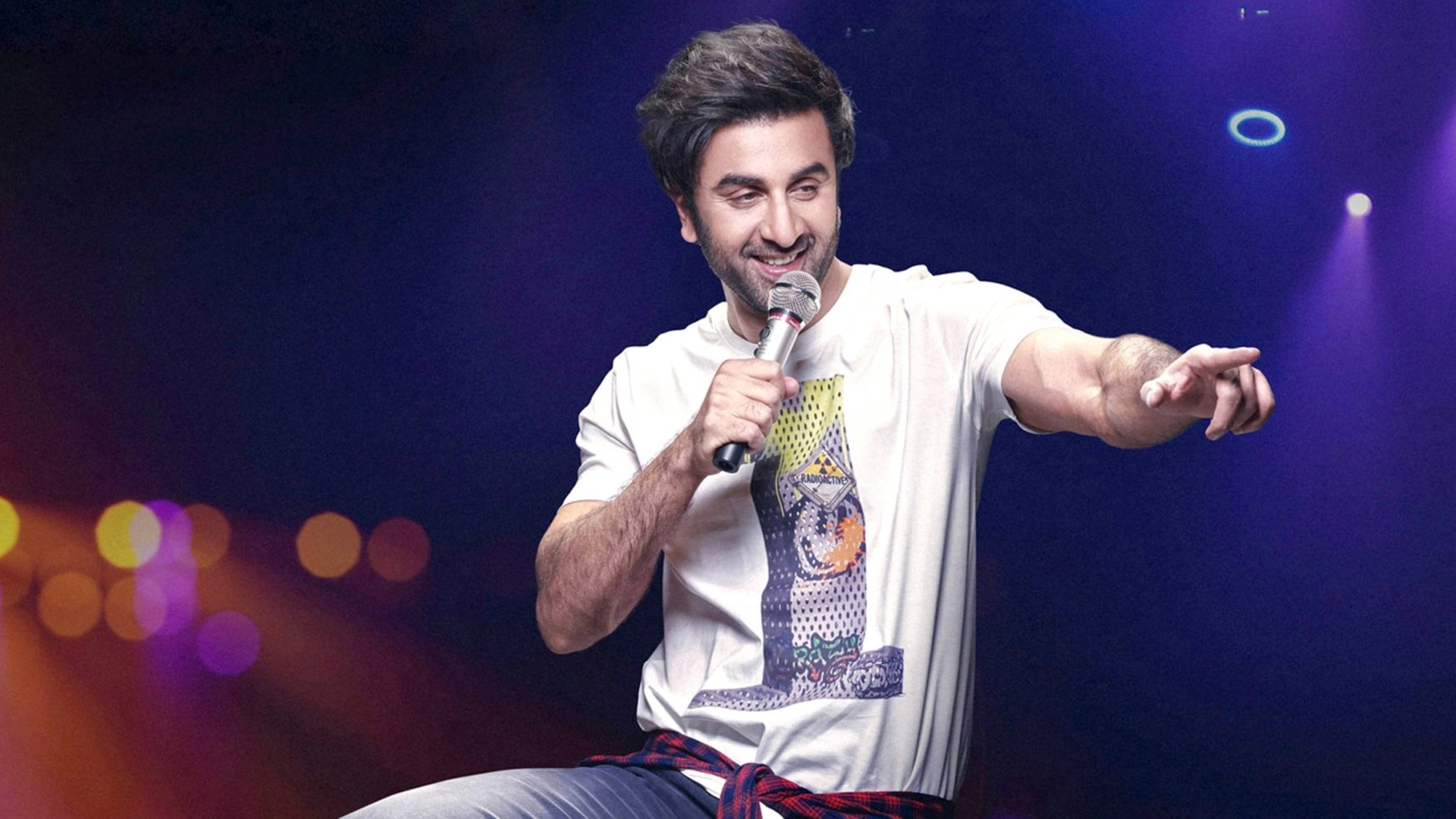 Ranbir Kapoor 桌布 壁紙 — 一個微笑的白T恤男子在色彩繽紛的舞台上，被鮮豔的聚光燈照亮，手持麥克風並做手勢。