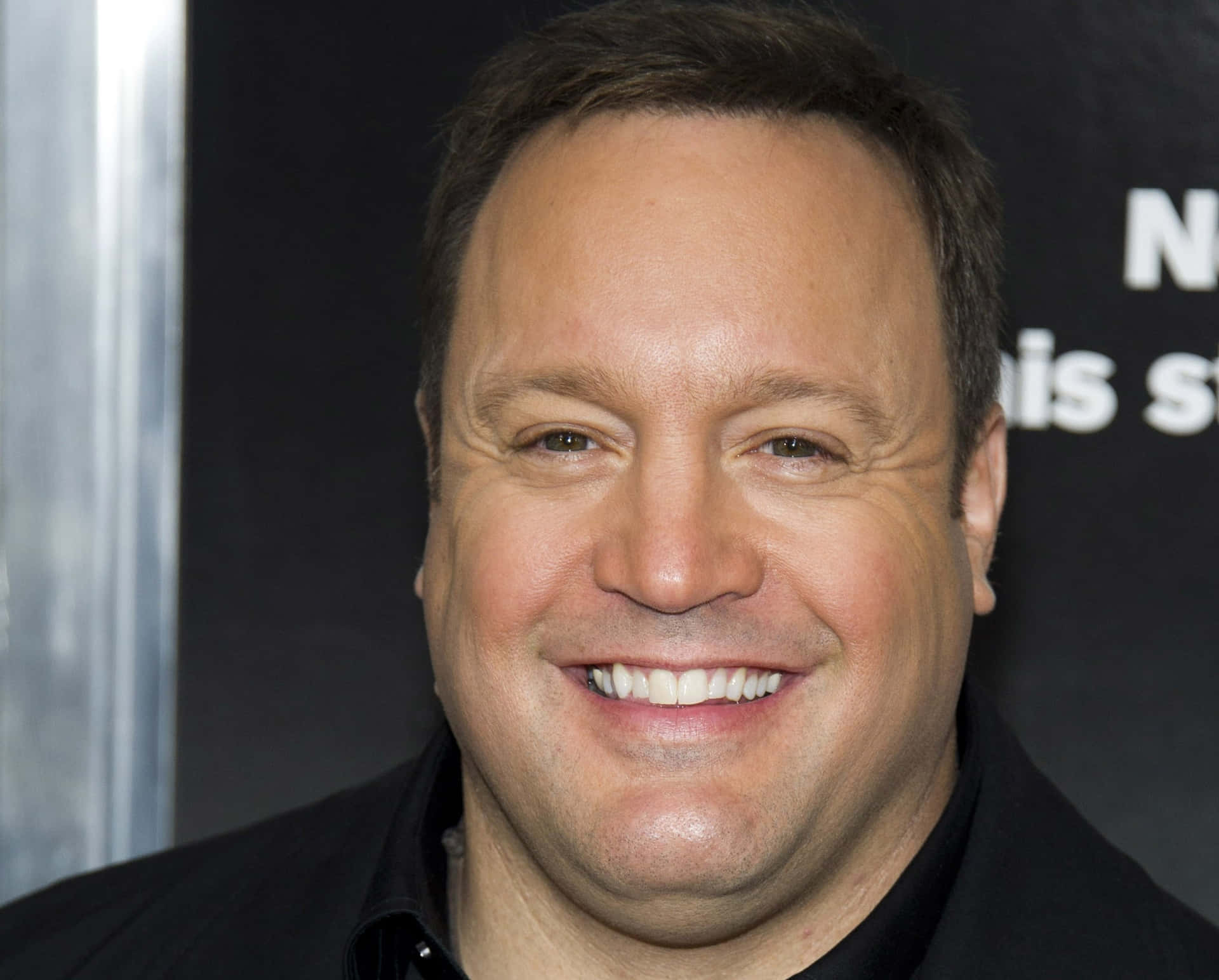 Kevin James 桌布 壁紙 — 一個微笑的短棕髮男人穿著黑色襯衫的特寫肖像，背景為深色並帶有部分白色文字。