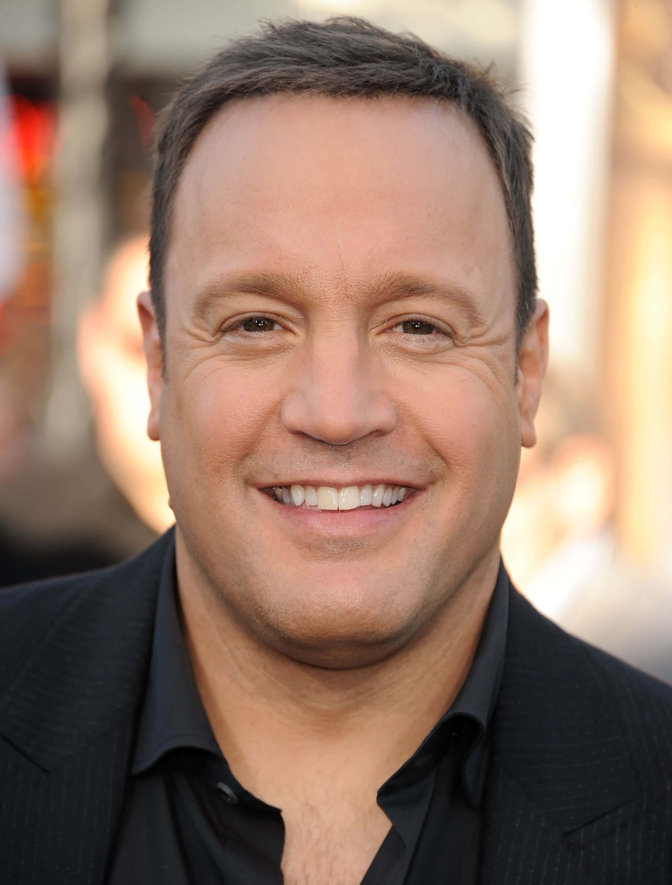 Kevin James 桌布 壁紙 — 一個微笑的短黑髮男人的特寫肖像，穿著黑色西裝和襯衫，背景是模糊的城市景觀。