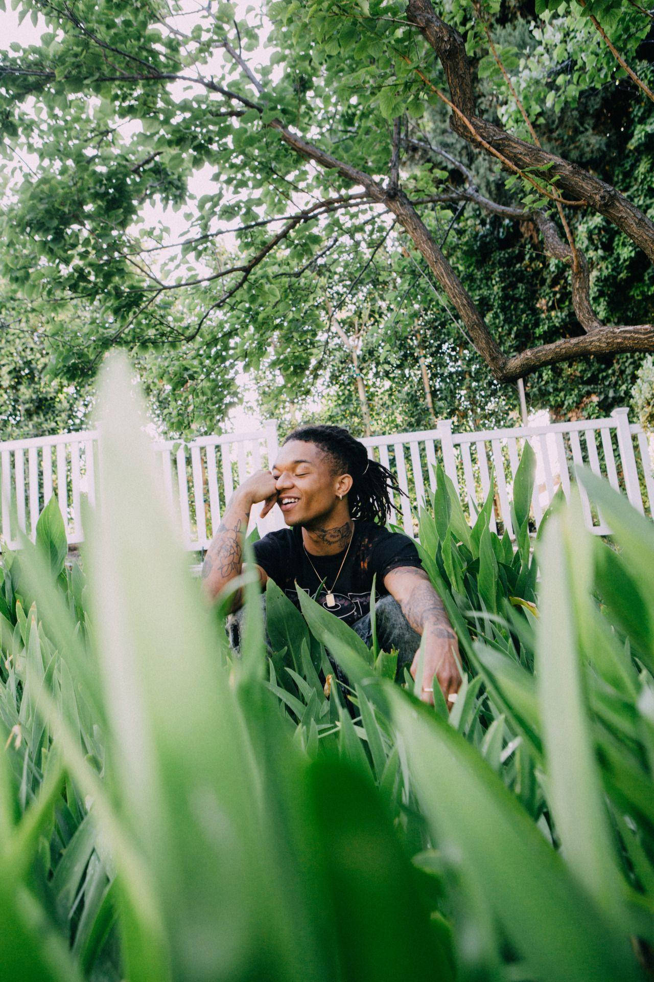 Swae Lee 桌布 壁紙 — 一個微笑的辮子男人坐在高大的綠色植物中，頭頂是枝葉茂密的樹木，身後有白色籬笆。