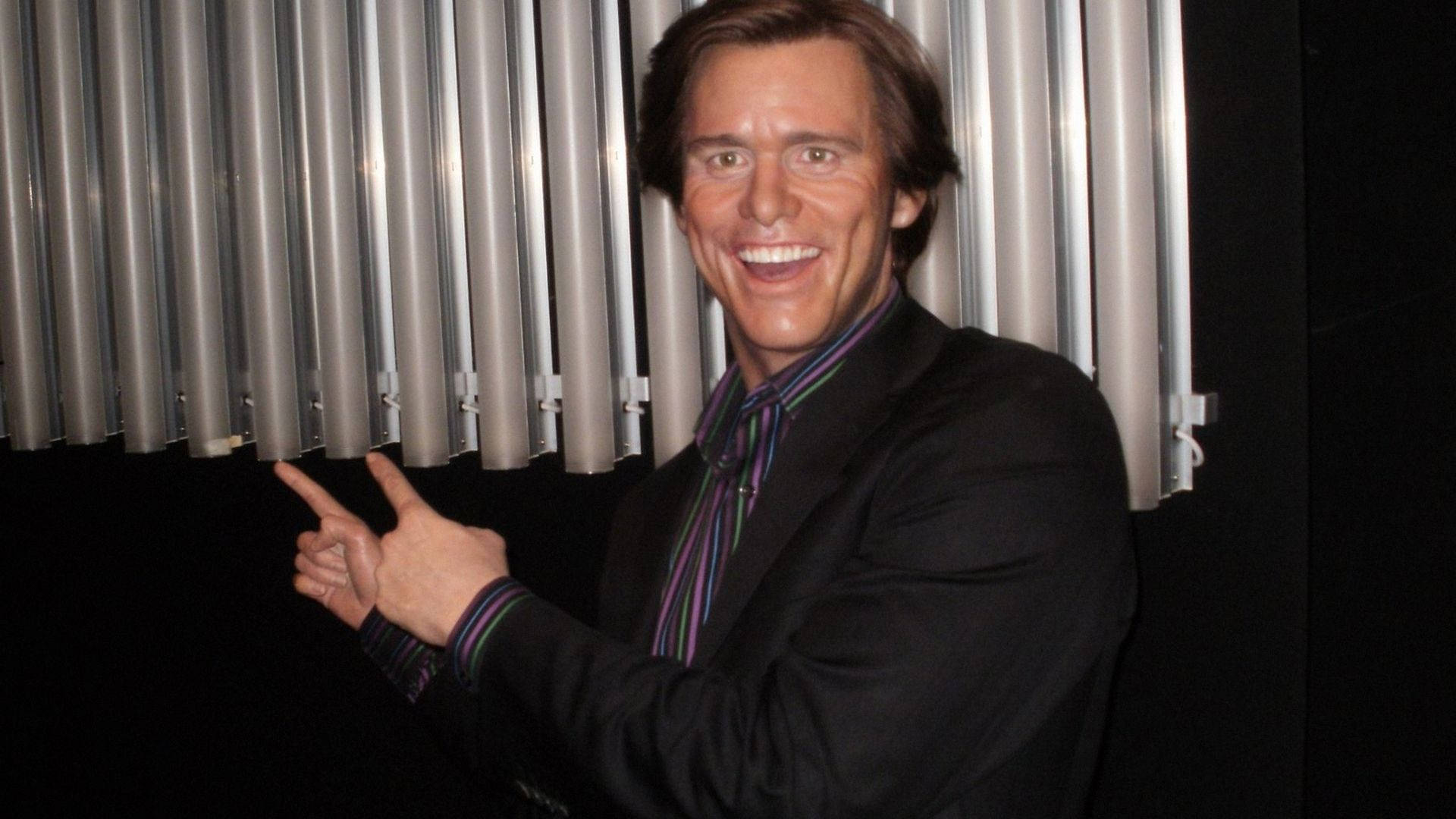 Jim Carrey 桌布 壁紙 — 一個微笑的金·凱瑞蠟像，身穿黑色西裝和條紋襯衫，在銀色垂直管道的背景下愉快地指著。