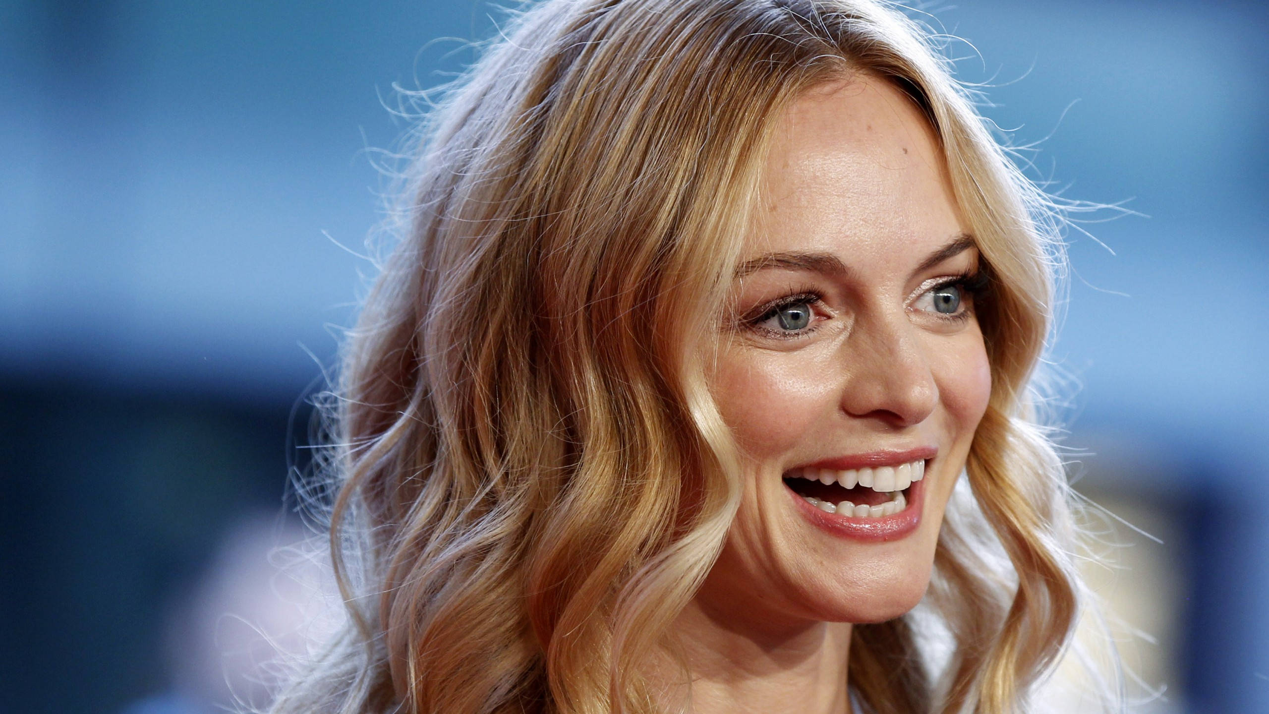 Heather Graham 桌布 壁紙 — 一個微笑的金髮女子的特寫肖像，擁有明亮的藍色眼睛和波浪狀頭髮，背景是模糊的藍色。