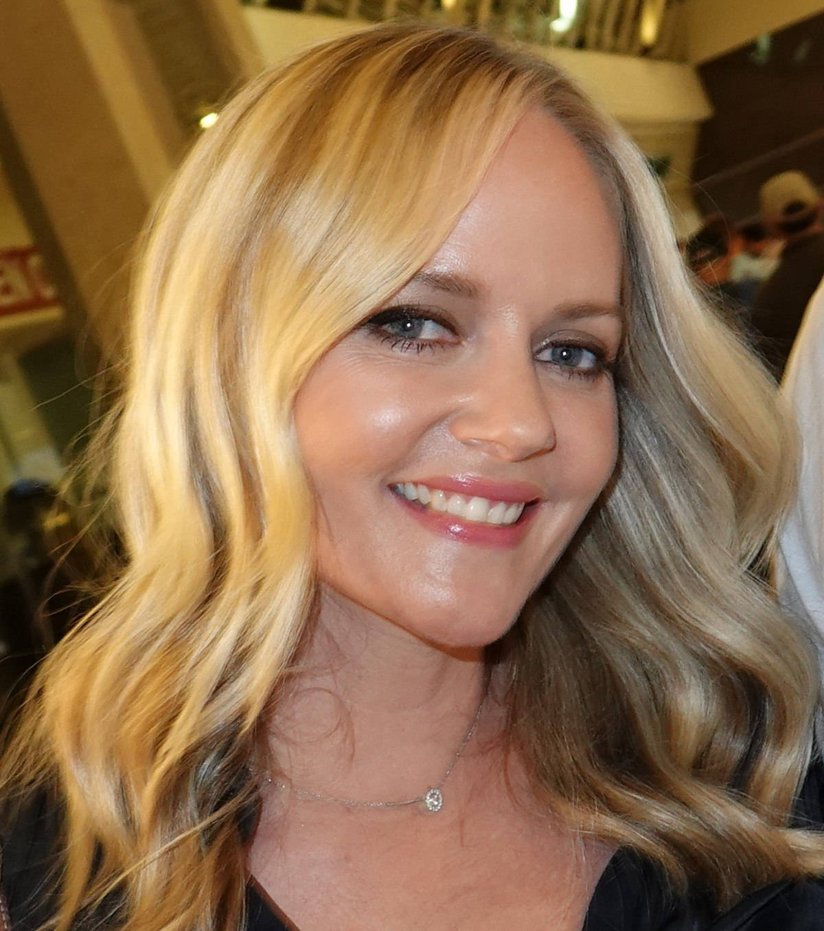 Celebrity 桌布 壁紙 — 一個微笑的金髮女性近距離肖像，波浪頭髮和精緻項鍊，在溫暖照明的室內環境中。