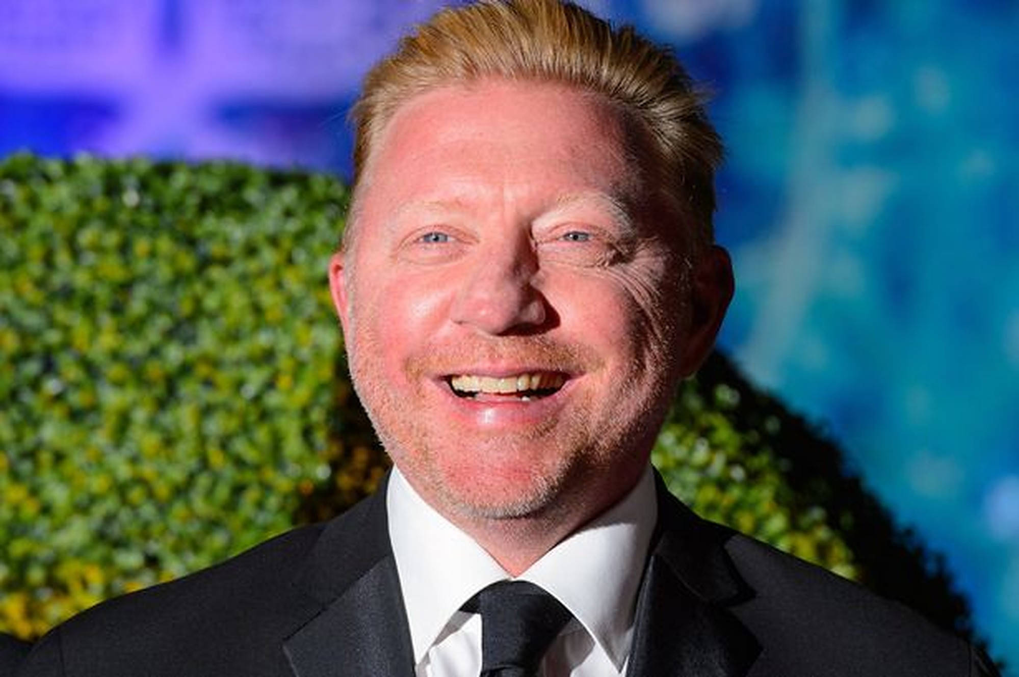 Boris Becker 桌布 壁紙 — 一個微笑的金髮男子，身穿黑色西裝，站在綠色籬笆和藍色背景前。