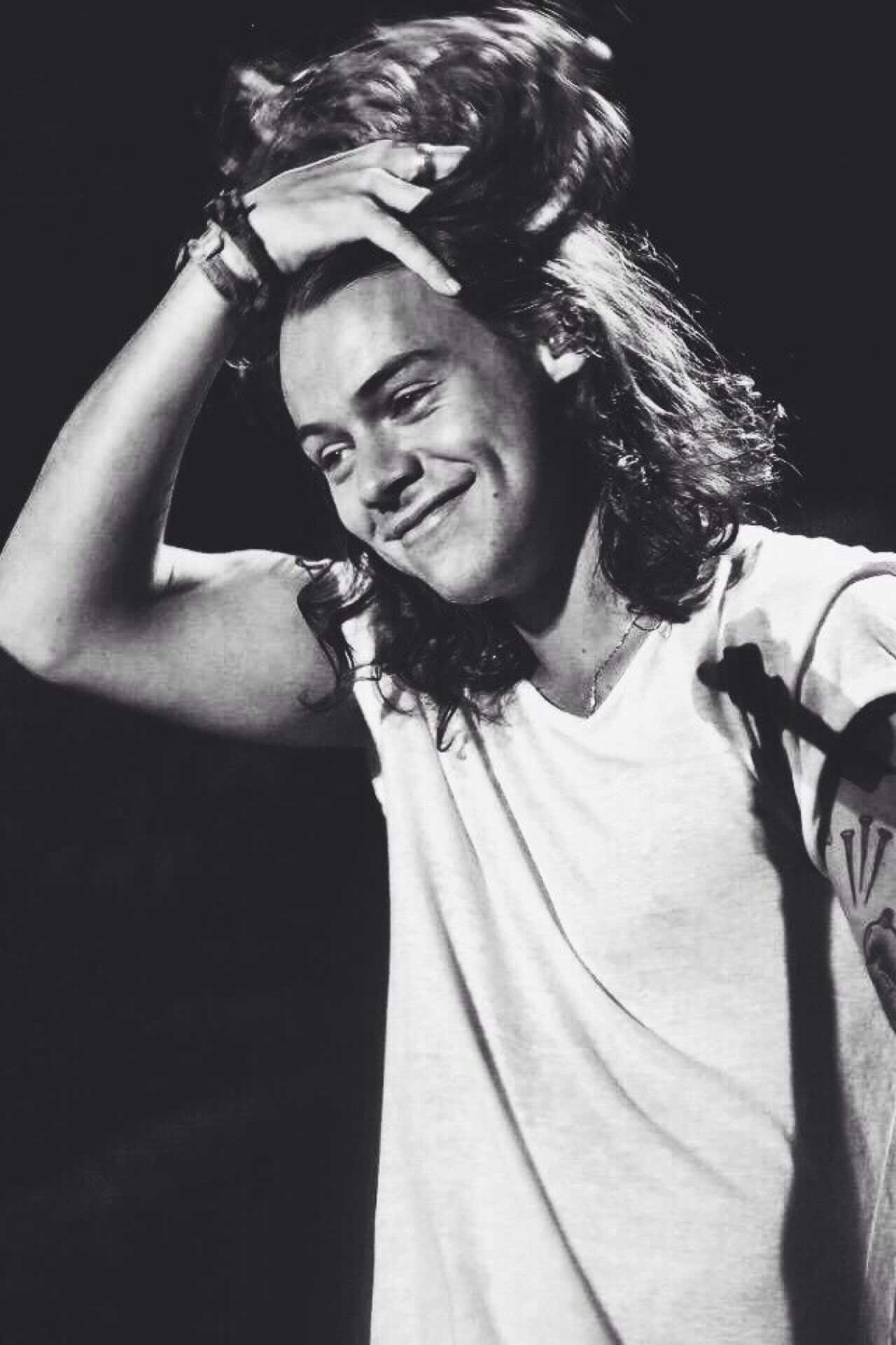 Harry Styles Black And White 桌布 壁紙 — 一個微笑的長髮年輕男子用手撥弄頭髮的黑白肖像。