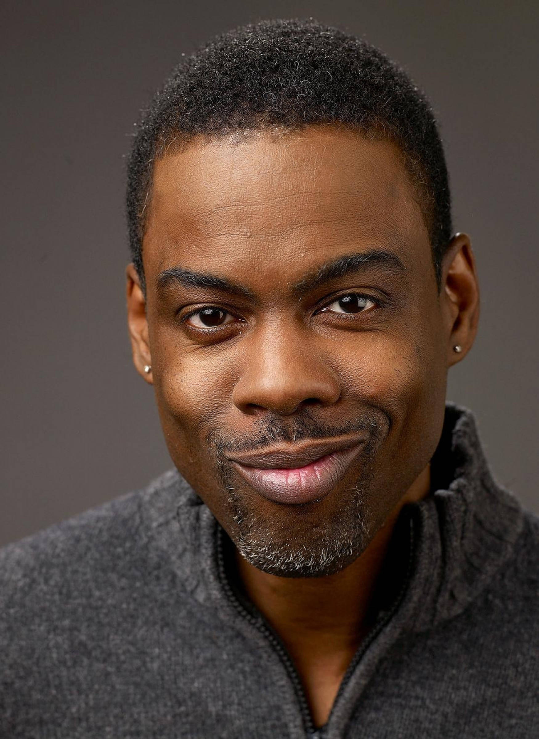 Chris Rock 桌布 壁紙 — 一個微笑的黑人男子特寫肖像，短髮和鬍渣，穿著灰色拉鍊領毛衣，背景為中性灰色。