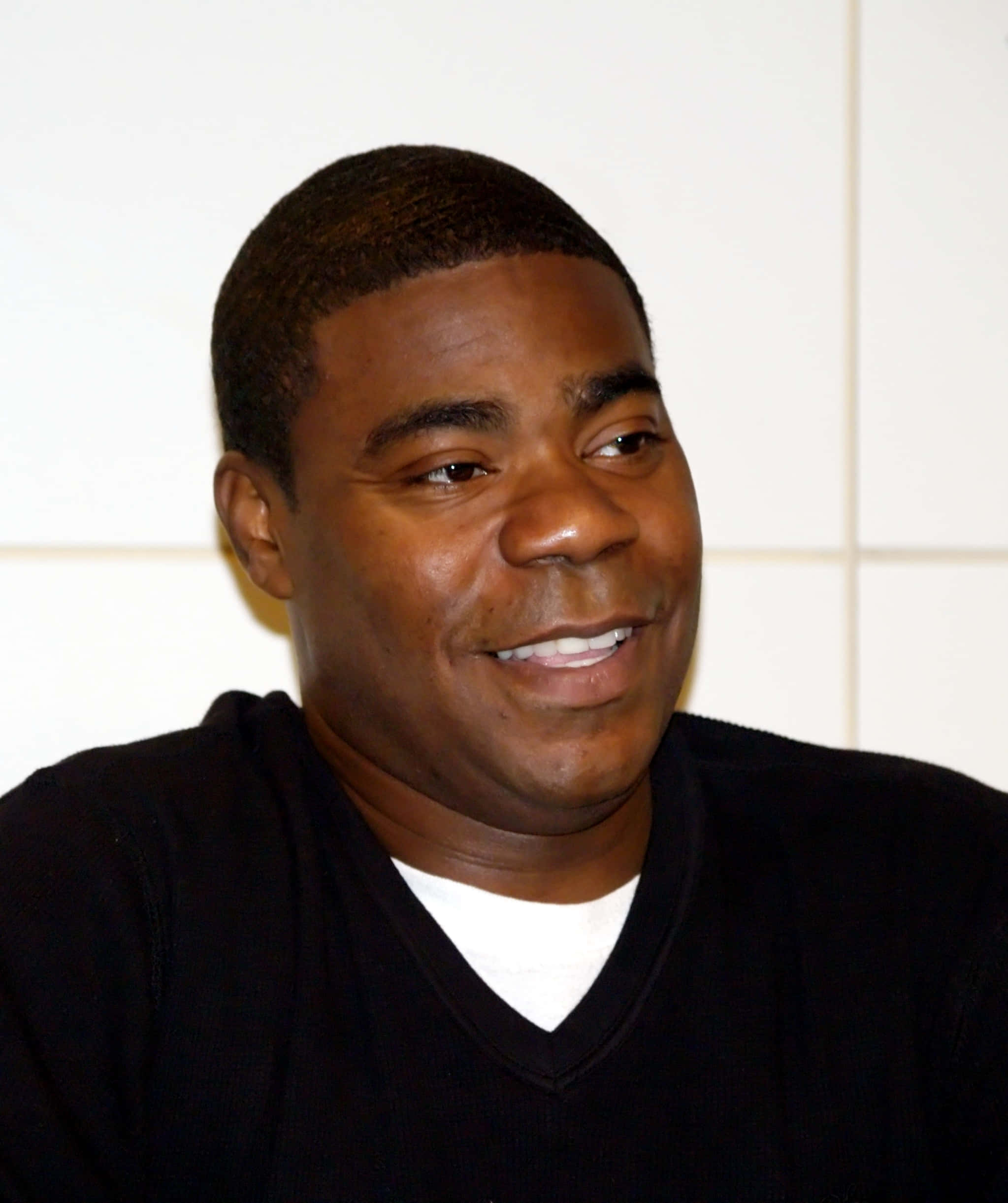 Tracy Morgan 桌布 壁紙 — 一個微笑的黑人男子身穿黑色毛衣內搭白色襯衫，背景是純白色瓷磚牆的頭像特寫。