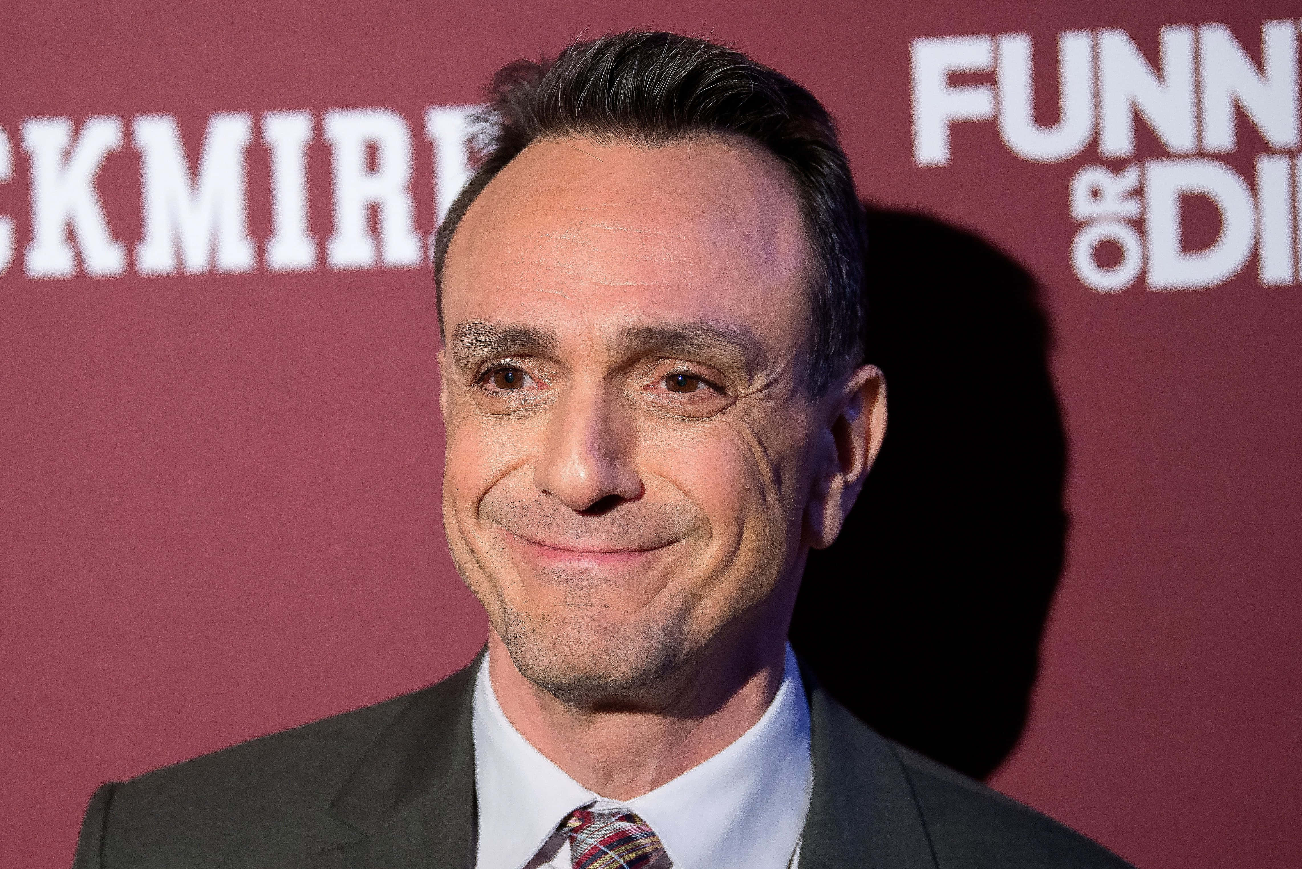 Hank Azaria 桌布 壁紙 — 一個微笑西裝男士的特寫肖像，背景為栗色，帶有白色文字。