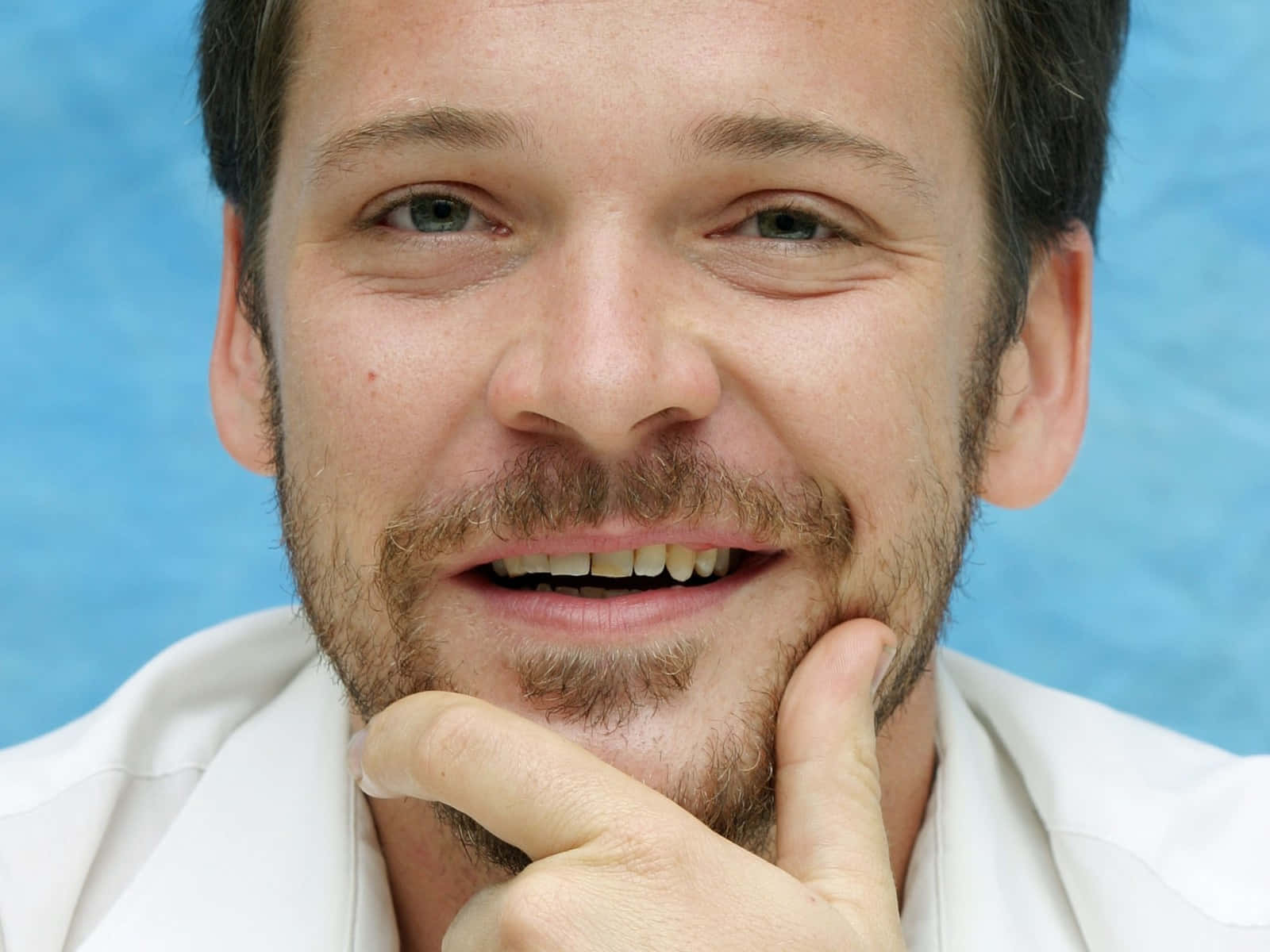 Peter Sarsgaard 桌布 壁紙 — 一個微笑鬍鬚男的近距離肖像，手放在下巴上，背景是藍色。