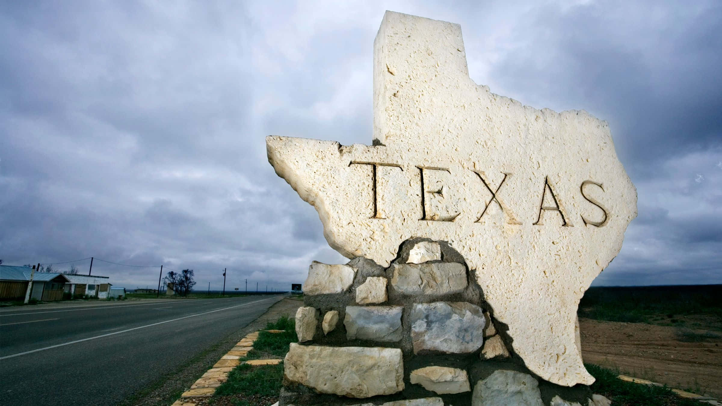 德州 桌布 壁紙 — 一個德州形狀的大型石製標誌，上面刻有「Texas」字樣，矗立在多雲天空下的路邊。