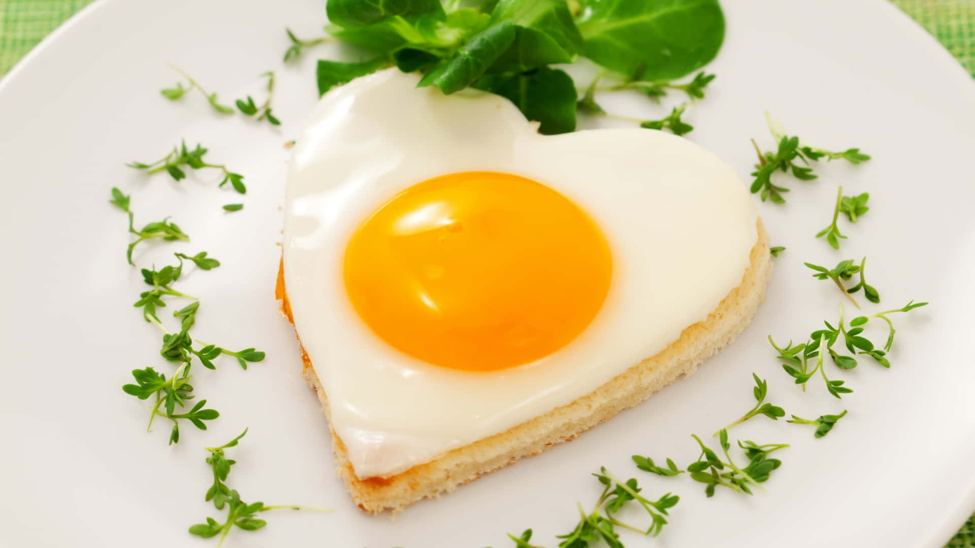 Egg White 桌布 壁紙 — 一個心形太陽蛋，蛋黃鮮豔，放在吐司上，配以新鮮綠色香草裝飾，置於白色盤子上。