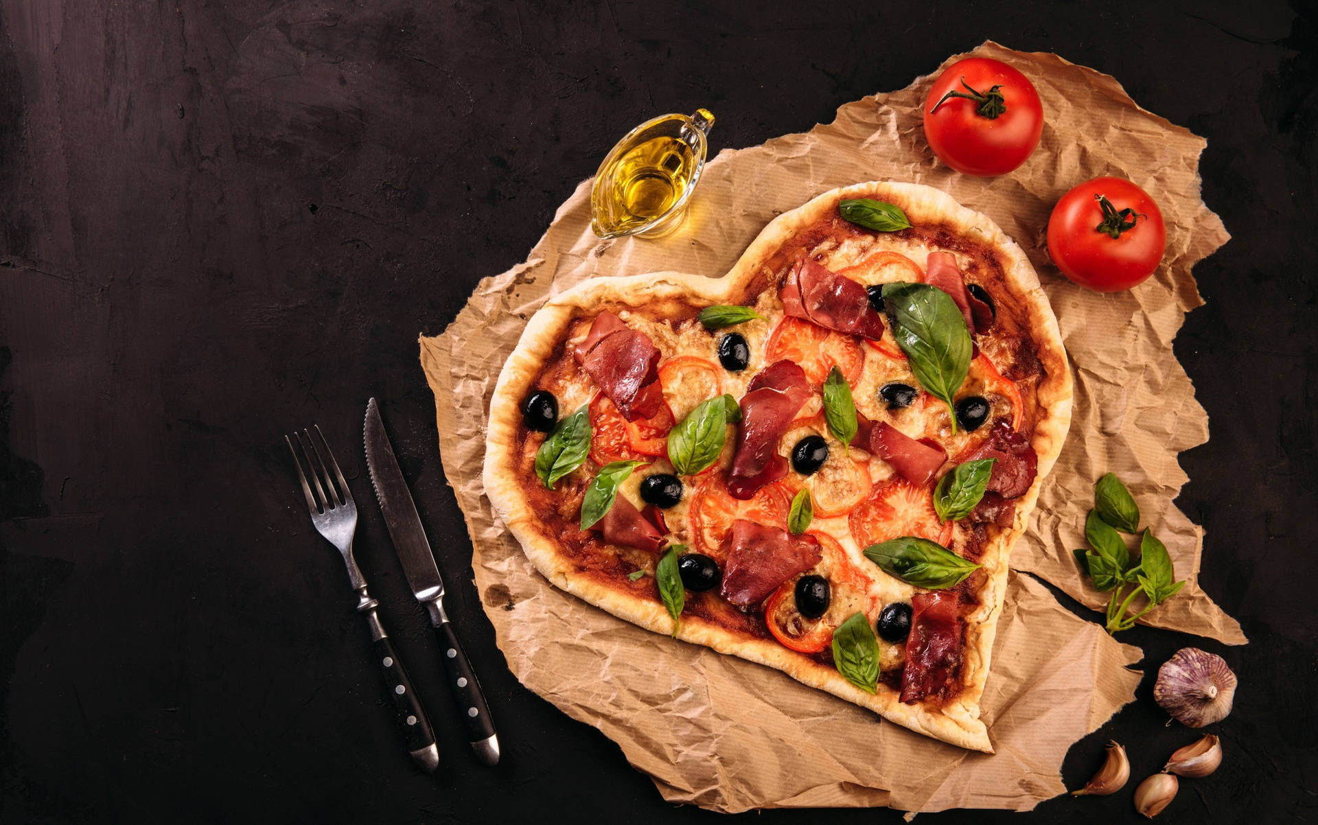 Pizza 桌布 壁紙 — 一個心形披薩，上面鋪滿羅勒、橄欖和火腿，放在皺巴巴的牛皮紙上，旁邊是新鮮番茄、大蒜和橄欖油。