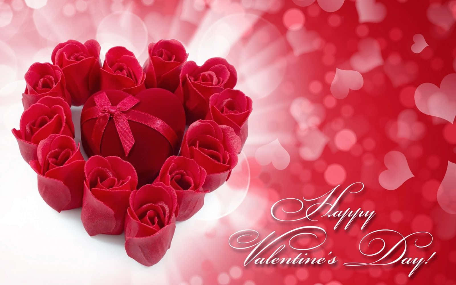 Valentines 桌布 壁紙 — 一個心形排列的紅玫瑰環繞著一個帶蝴蝶結的紅色禮物盒，背景是發光的紅色散景，有漂浮的心形和'Happy Valentine's Day'字樣。