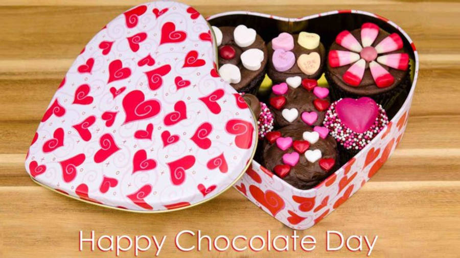 Chocolate Day 桌布 壁紙 — 一個心形盒子裝滿了巧克力杯子蛋糕，上面有粉紅色、白色和紅色的心形糖果，放在木質表面上。