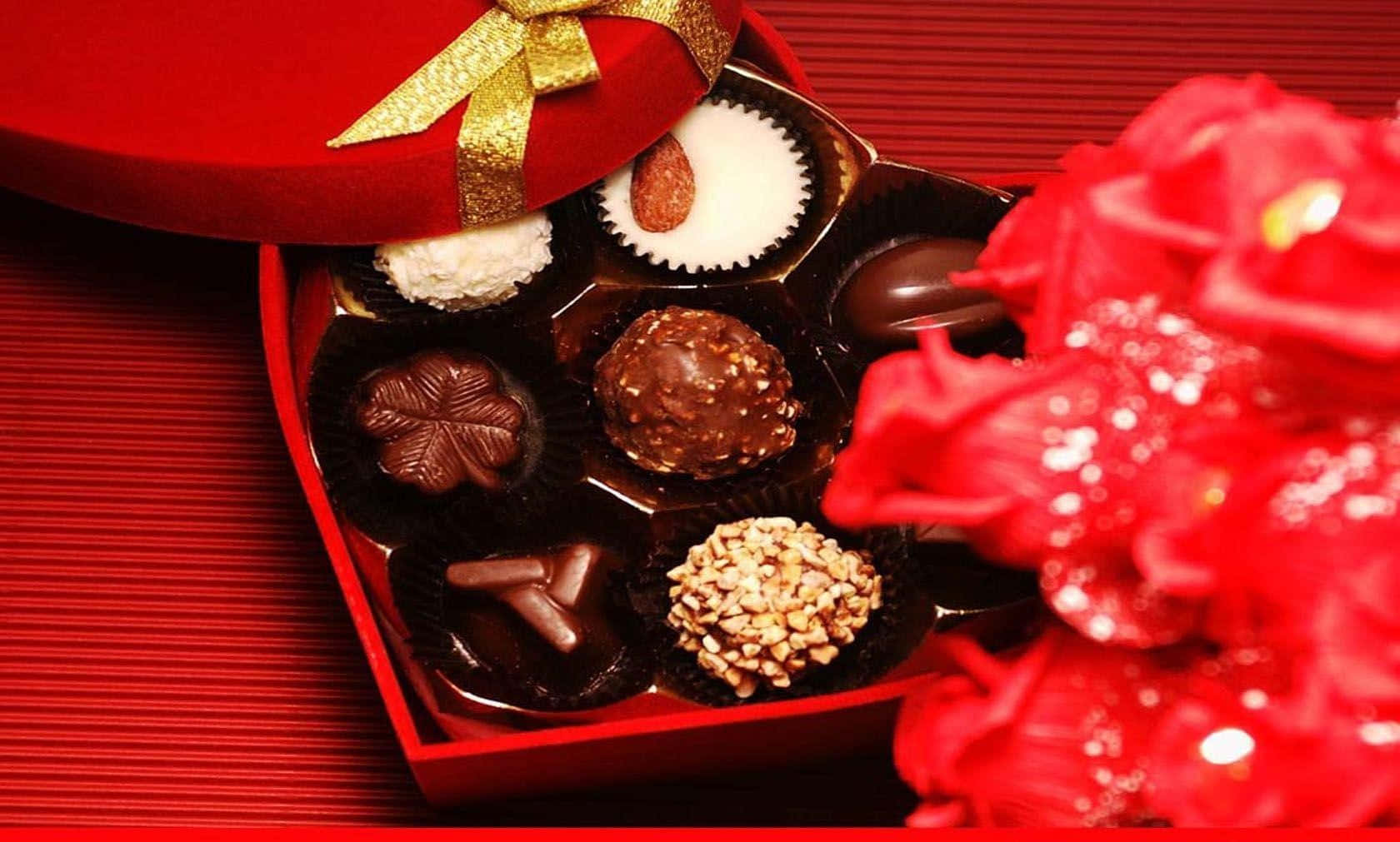 Chocolate 桌布 壁紙 — 一個心形紅色綜合巧克力盒放在紋理紅色表面上，旁邊是鮮豔的紅玫瑰。