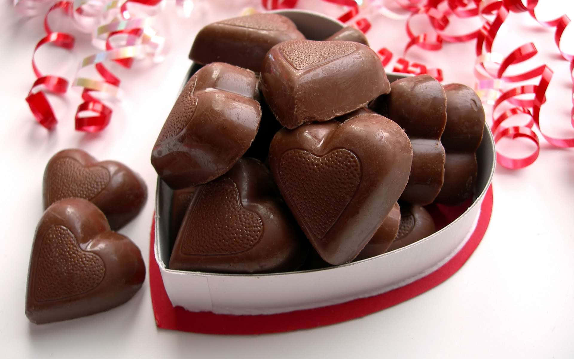 Brown Chocolate 桌布 壁紙 — 一個心形金屬盒，裝滿了閃亮的黑巧克力心形糖果，周圍環繞著紅色絲帶卷。