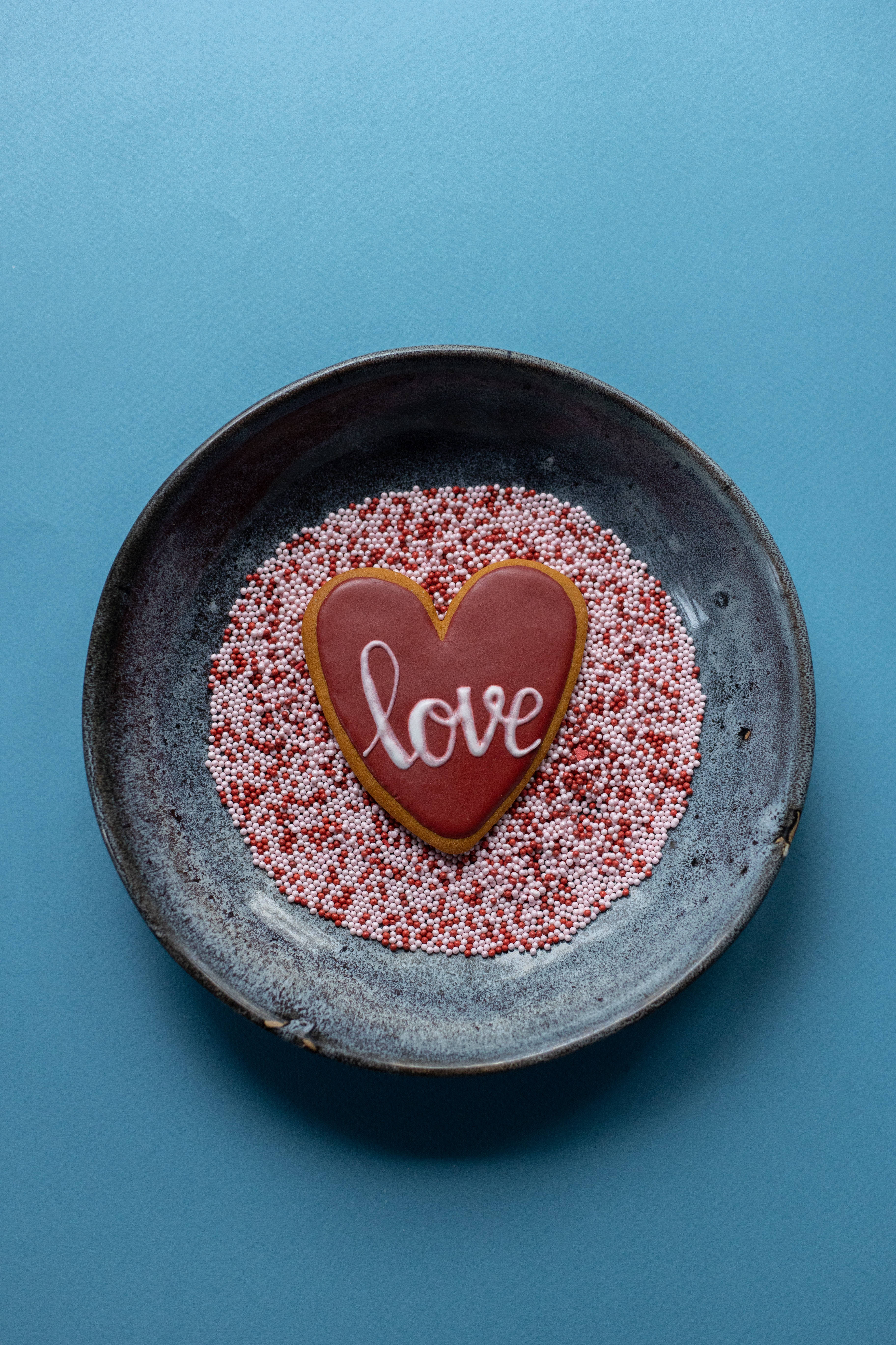 Cute Blue 桌布 壁紙 — 一個心形餅乾，上面用白色糖霜寫著「love」，放在紅色和白色糖粒上，置於一個質樸的陶瓷碗中，背景為藍色。