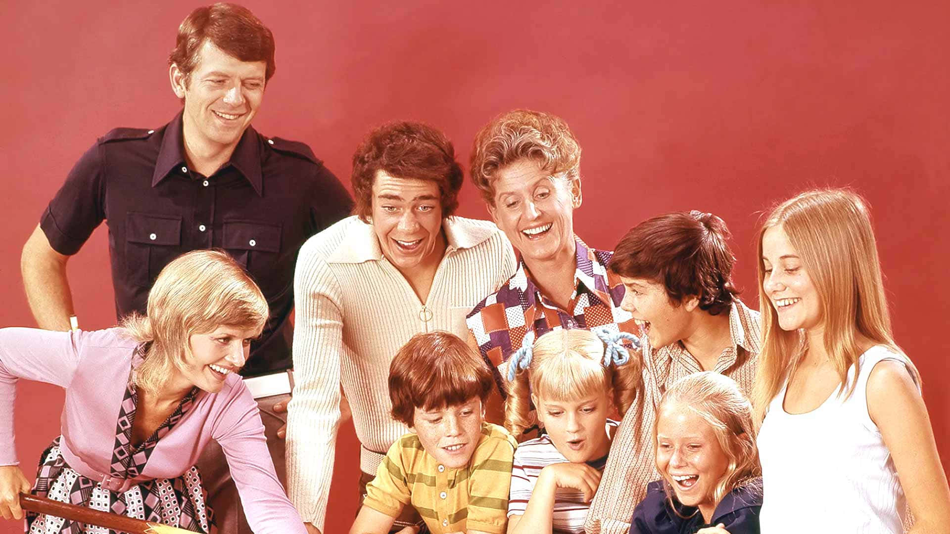 show 桌布 壁紙 — 一個快樂的1970年代家庭群體在純紅色背景前一起擺姿勢，帶著燦爛的笑容和復古服裝。