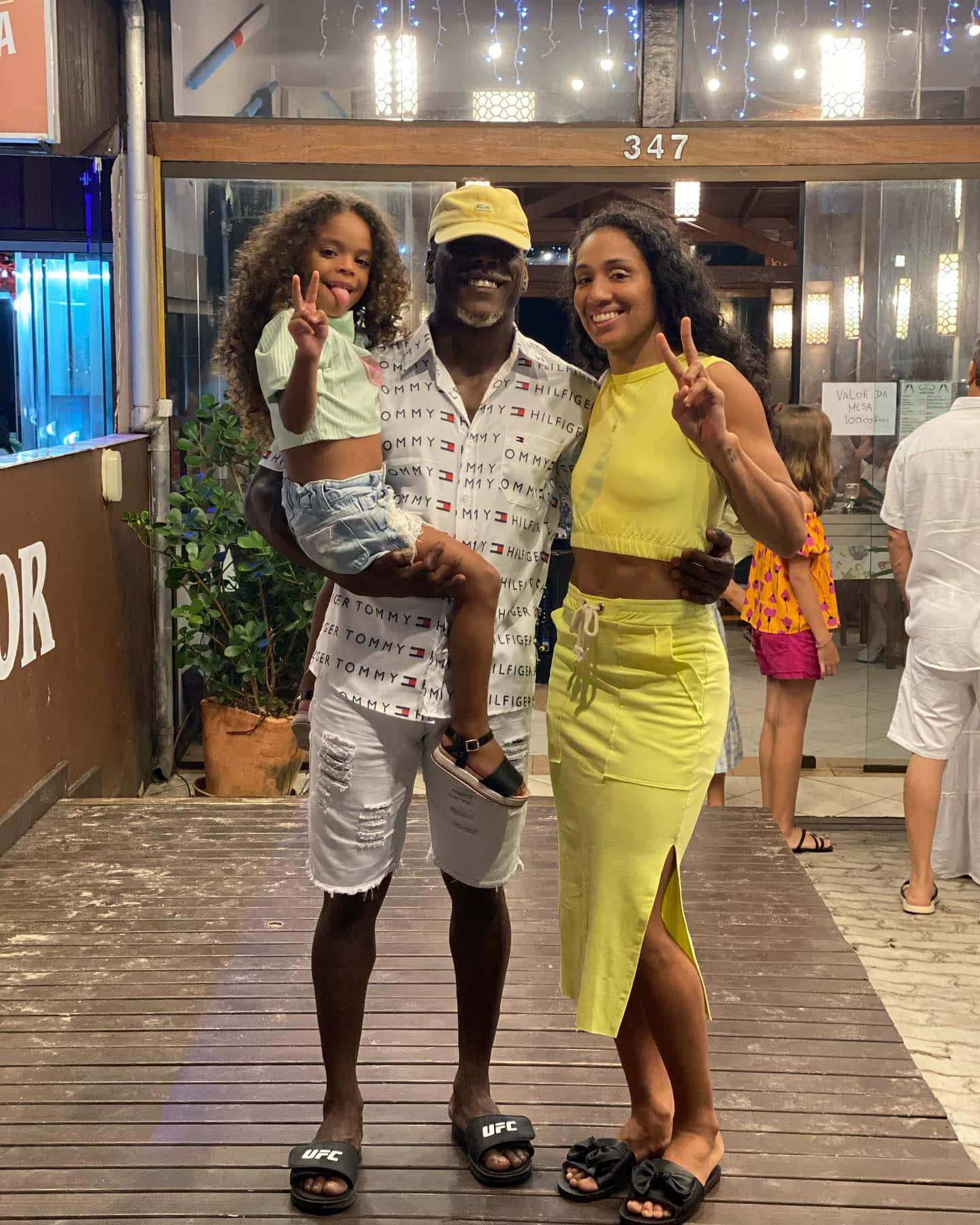 Family 桌布 壁紙 — 一個快樂的三口之家在夜間的木甲板上拍照，身後有燈串和商店門面。