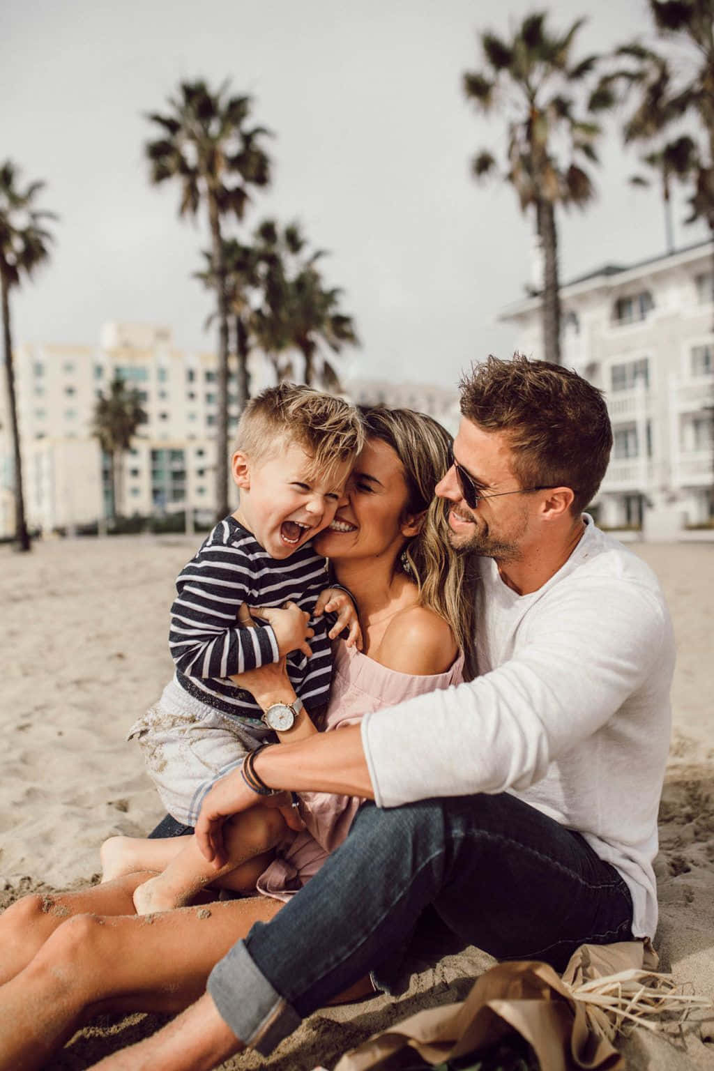 Family Beach 桌布 壁紙 — 一個快樂的三口之家在沙灘上一起大笑，背景有棕櫚樹和海濱建築。