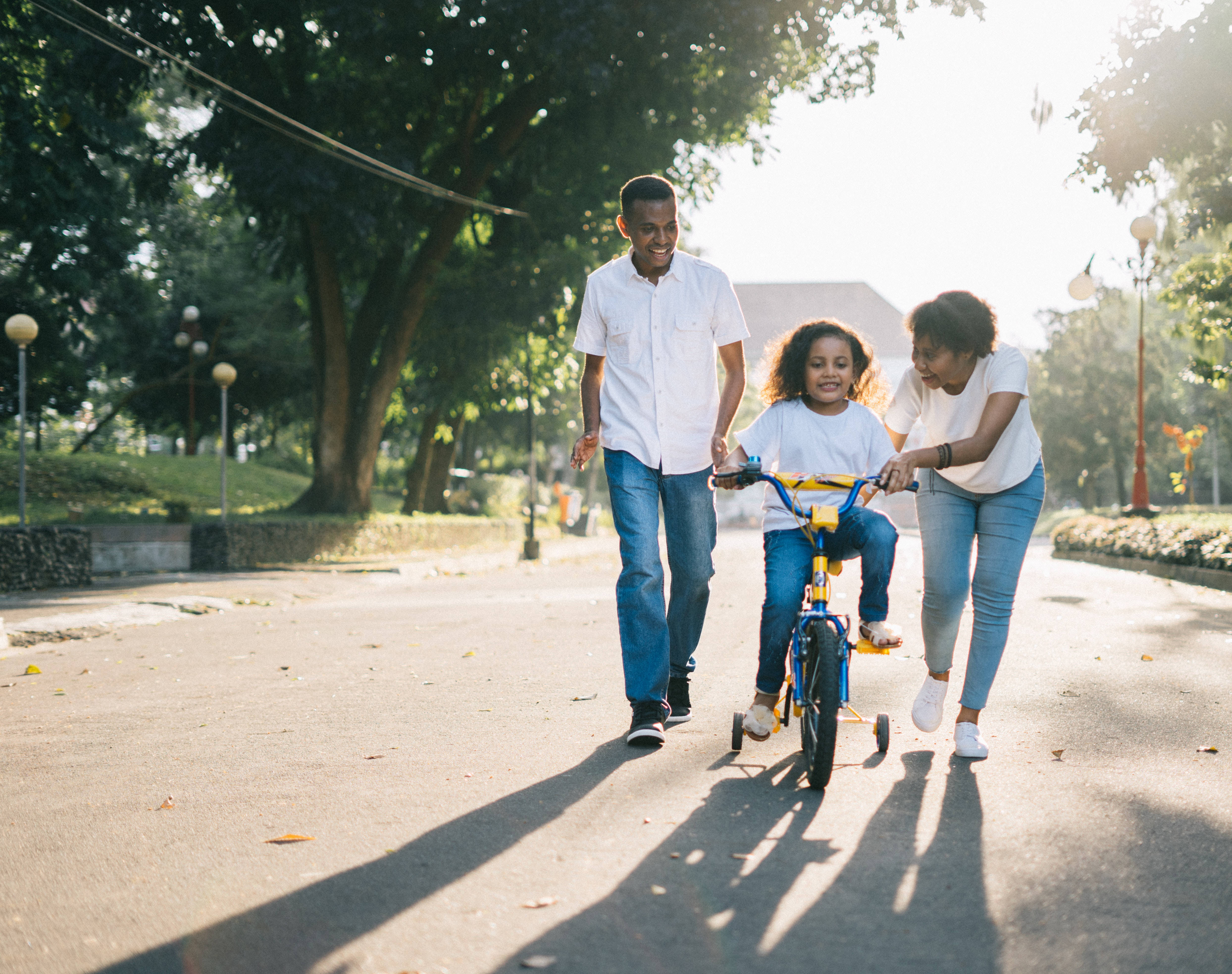 Family 桌布 壁紙 — 一個快樂的三口之家在陽光明媚的公園裡一起散步，孩子騎著三輪車，父母在旁支持。