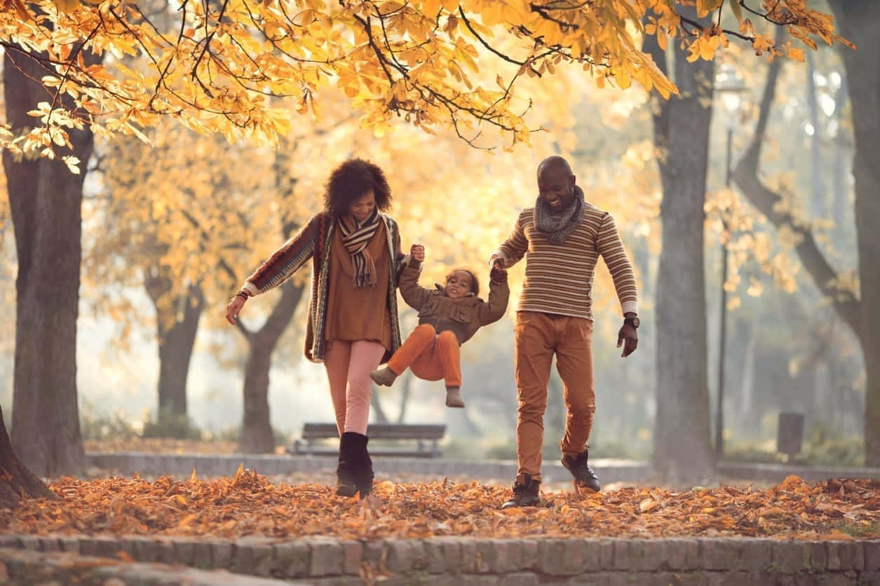 Fall Family 桌布 壁紙 — 一個快樂的三口之家在陽光明媚的秋日公園裡散步，落葉在金色樹冠下圍繞他們的腳邊旋轉。
