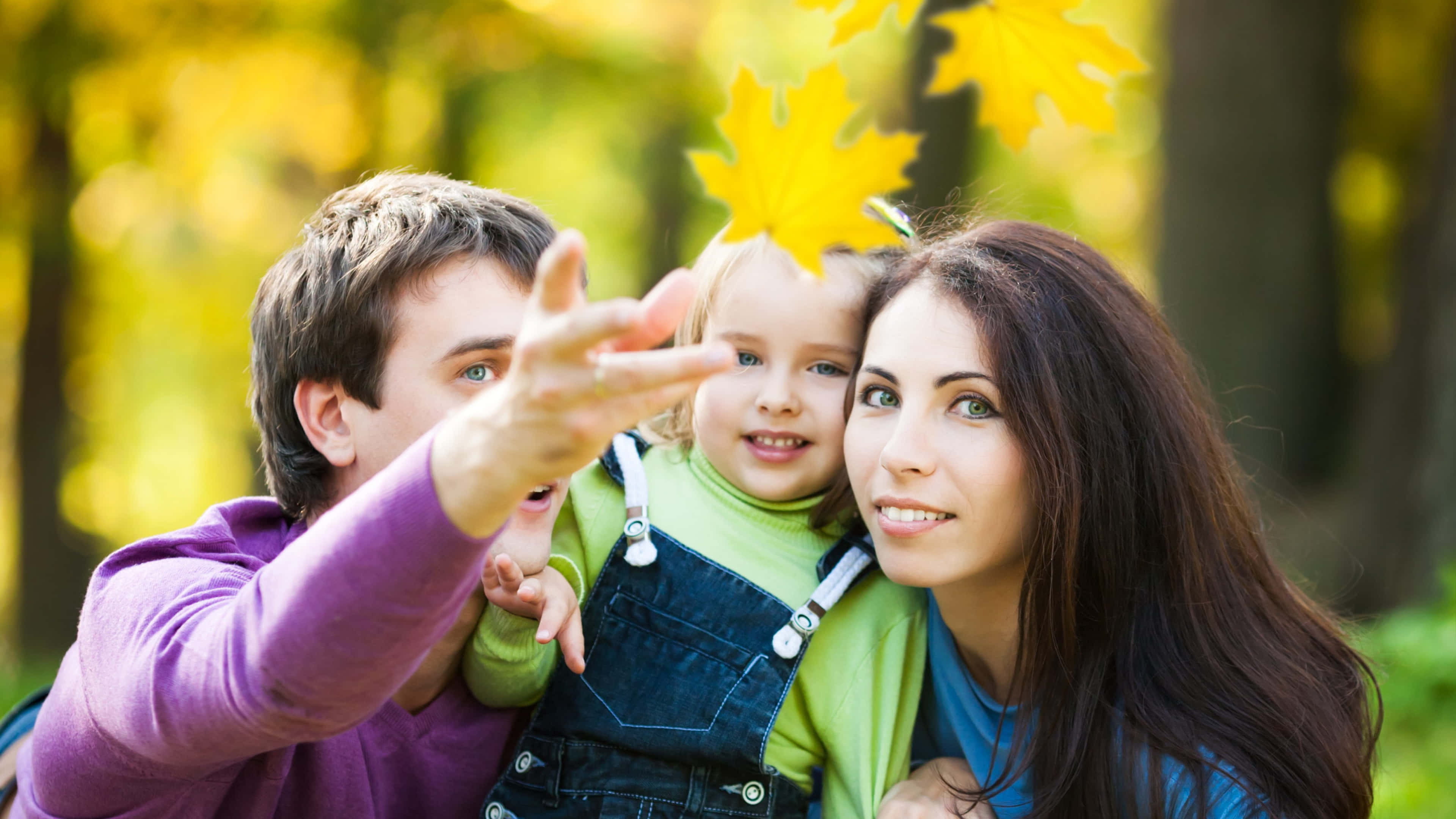 Family 桌布 壁紙 — 一個快樂的三口之家在陽光明媚的秋日森林中一起大笑，孩子拿著一片鮮黃色的楓葉。