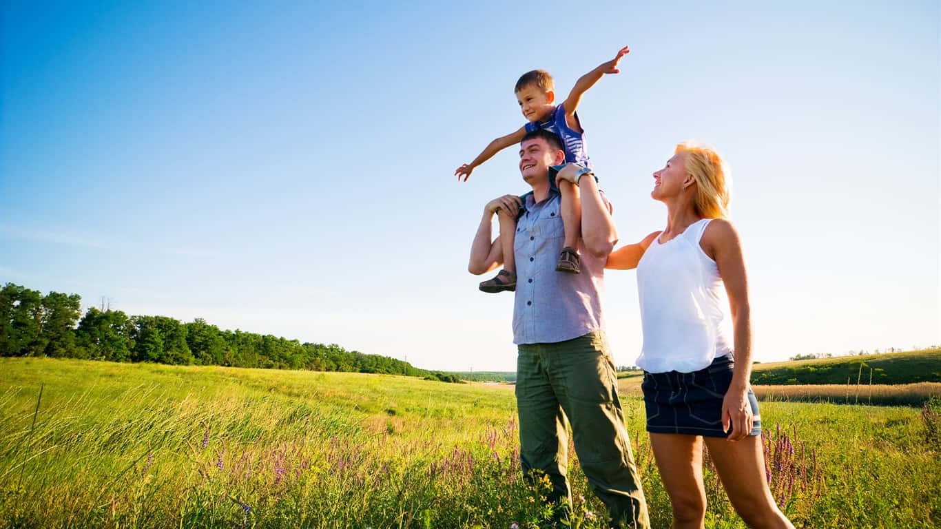 Family 桌布 壁紙 — 一個快樂的三口之家在陽光明媚的草地上大笑，周圍有高高的草和遠處的樹木，頭頂是清澈的藍天。