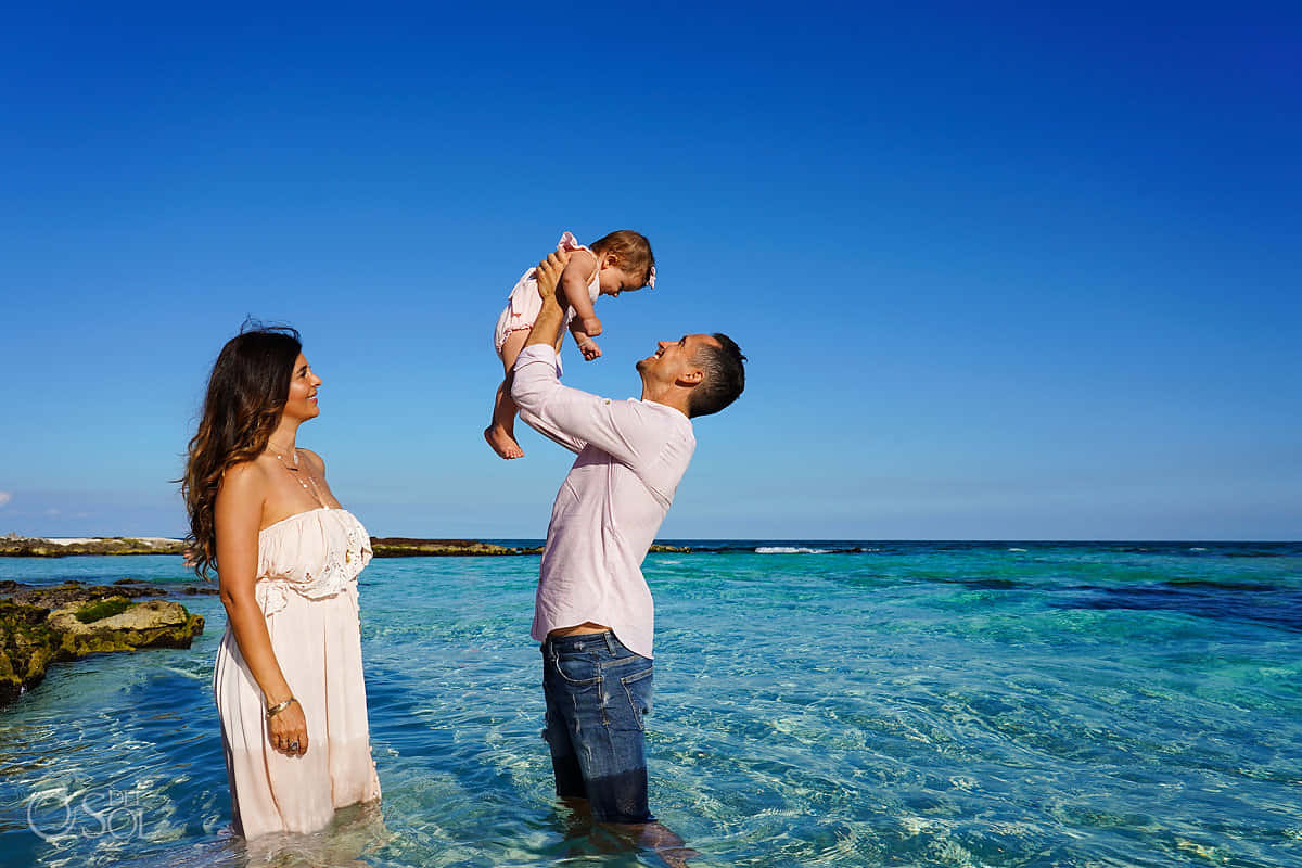 Family Portrait 桌布 壁紙 — 一個快樂的三口之家站在清澈的碧綠海水中，頭頂廣闊藍天，男人將他們的寶寶高高舉起，女人在旁邊微笑。