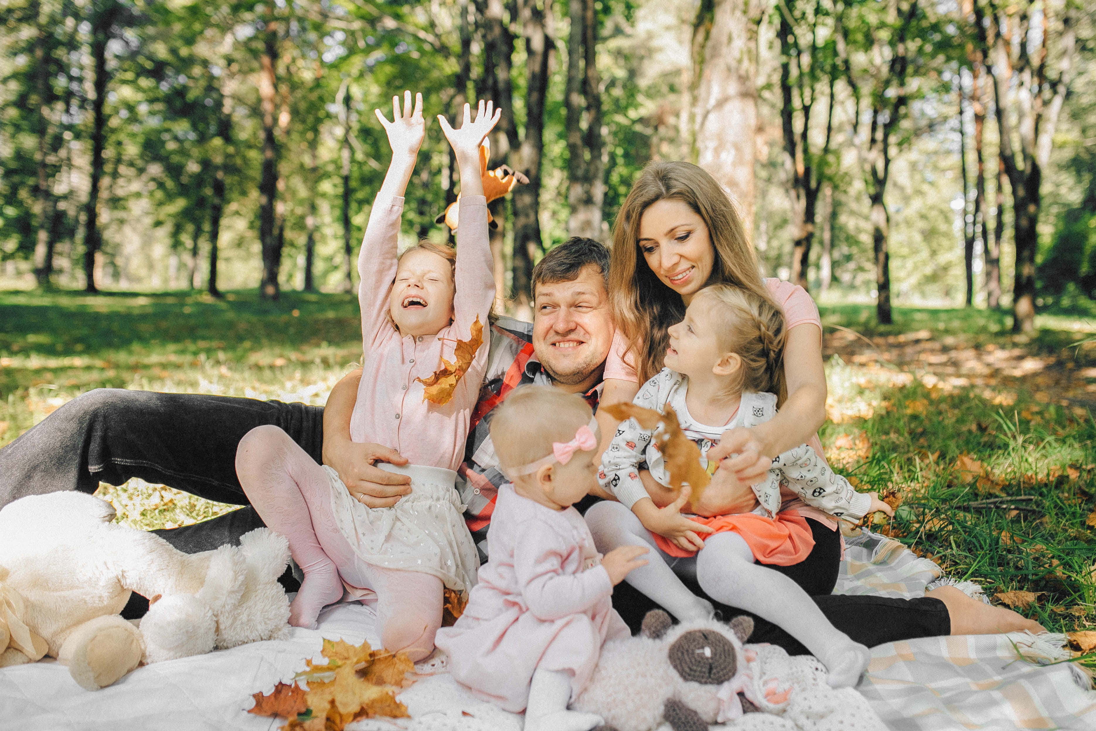 Fall Family 桌布 壁紙 — 一個快樂的五口之家在陽光明媚的秋日森林中，在野餐墊上笑著玩絨毛玩具。