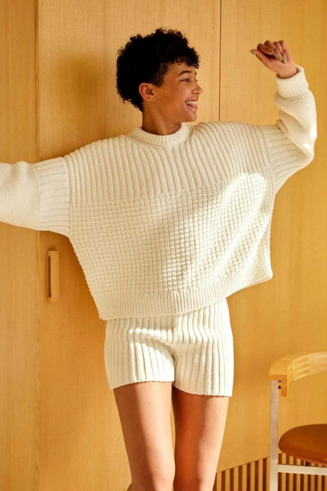 Minimalist Fashion 桌布 壁紙 — 一個快樂的人穿著奶油色針織毛衣和短褲，站在溫暖的木牆前，雙臂張開。