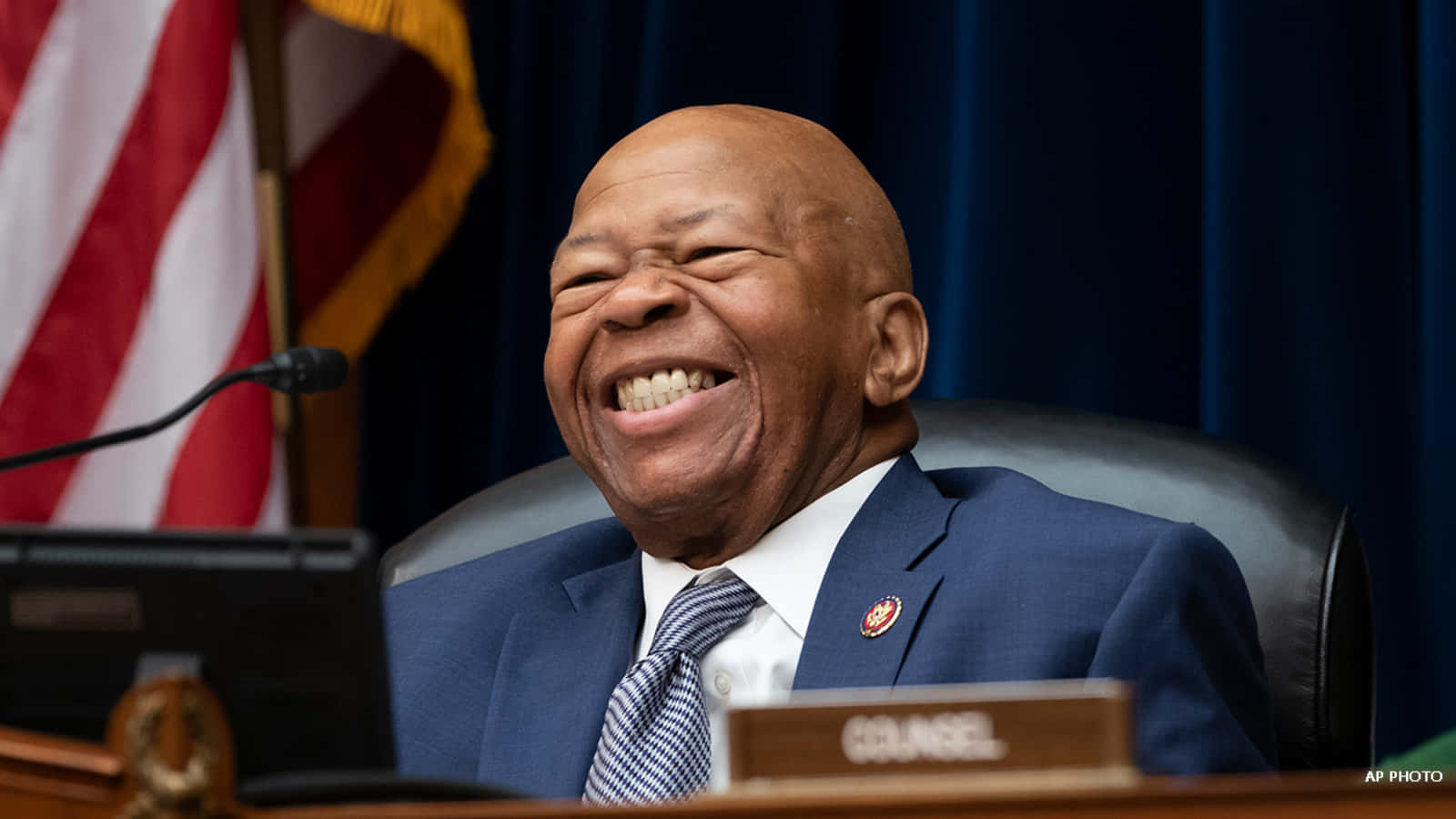 smiling 桌布 壁紙 — 一個快樂的光頭男人穿著藍色西裝，坐在木桌旁，身後有美國國旗，開懷大笑。