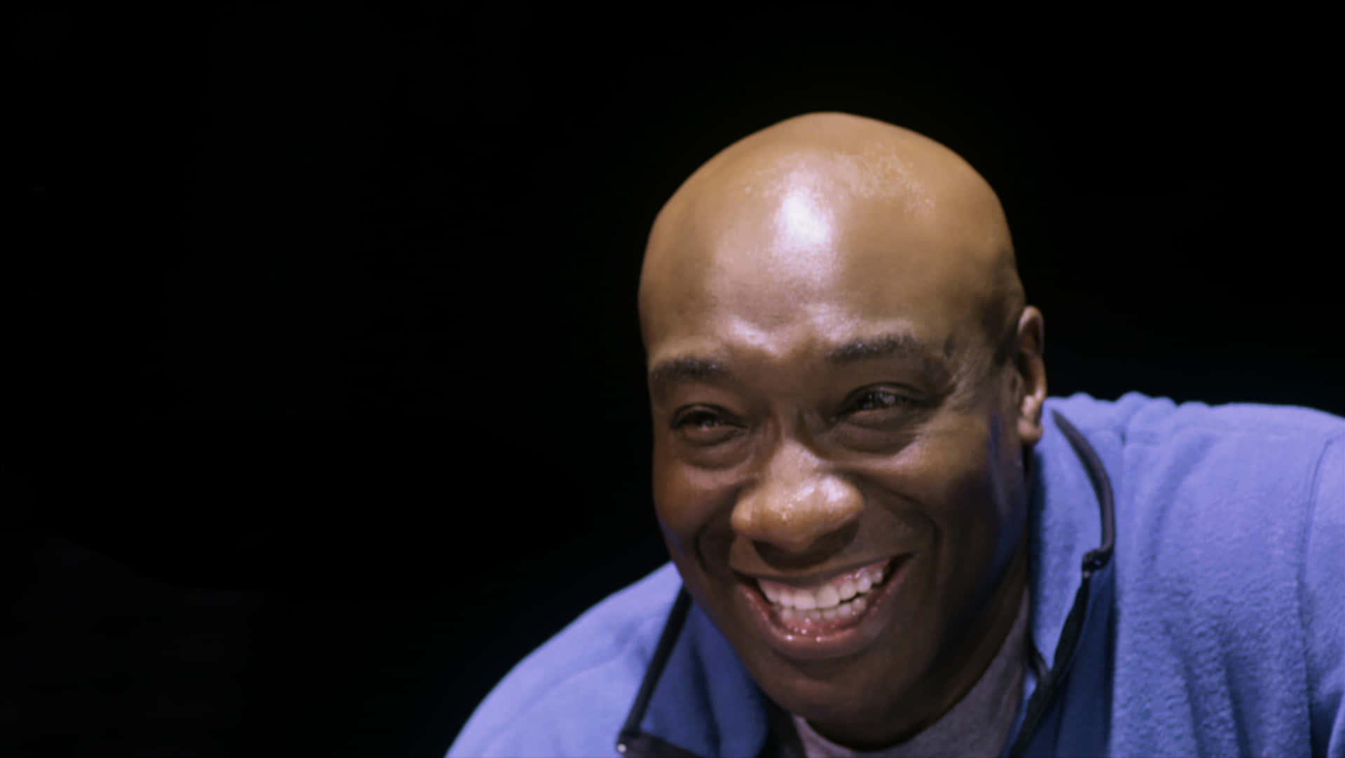 Michael Clarke Duncan 桌布 壁紙 — 一個快樂的光頭男子穿著藍色連帽衫，在純黑背景上露出燦爛的笑容。