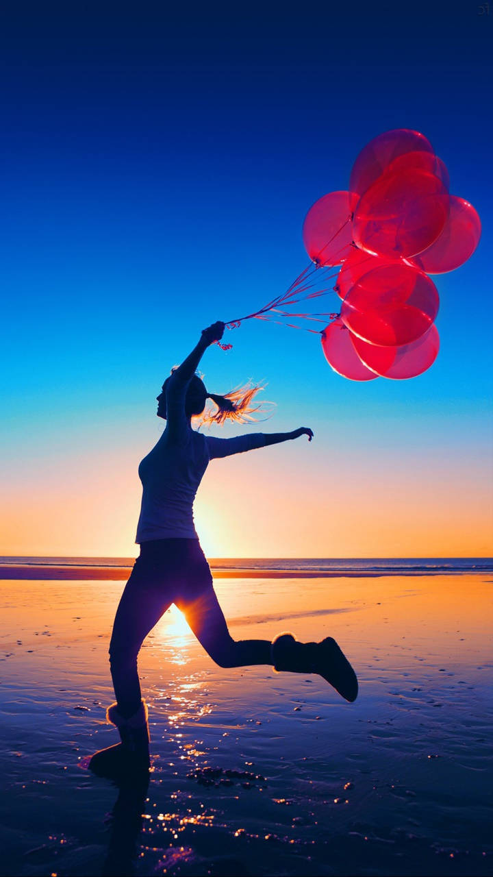 Balloon 桌布 壁紙 — 一個快樂的剪影在日落時分的沙灘上跳躍，手持發光的紅色氣球，背景是漸層的天空。