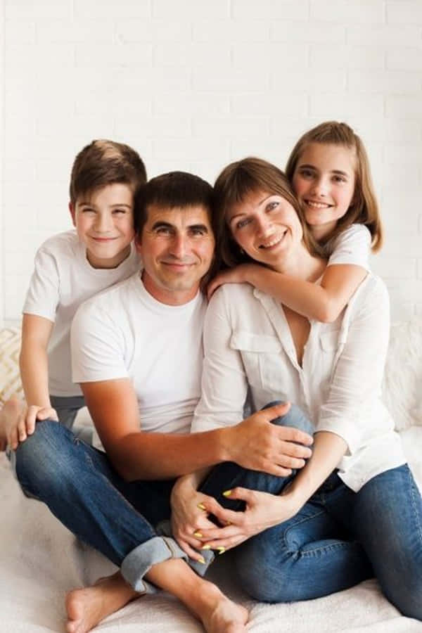 Family Portrait 桌布 壁紙 — 一個快樂的四口之家，身穿白色襯衫和牛仔褲，在白色磚牆前的床上緊密地坐在一起，溫暖地微笑。