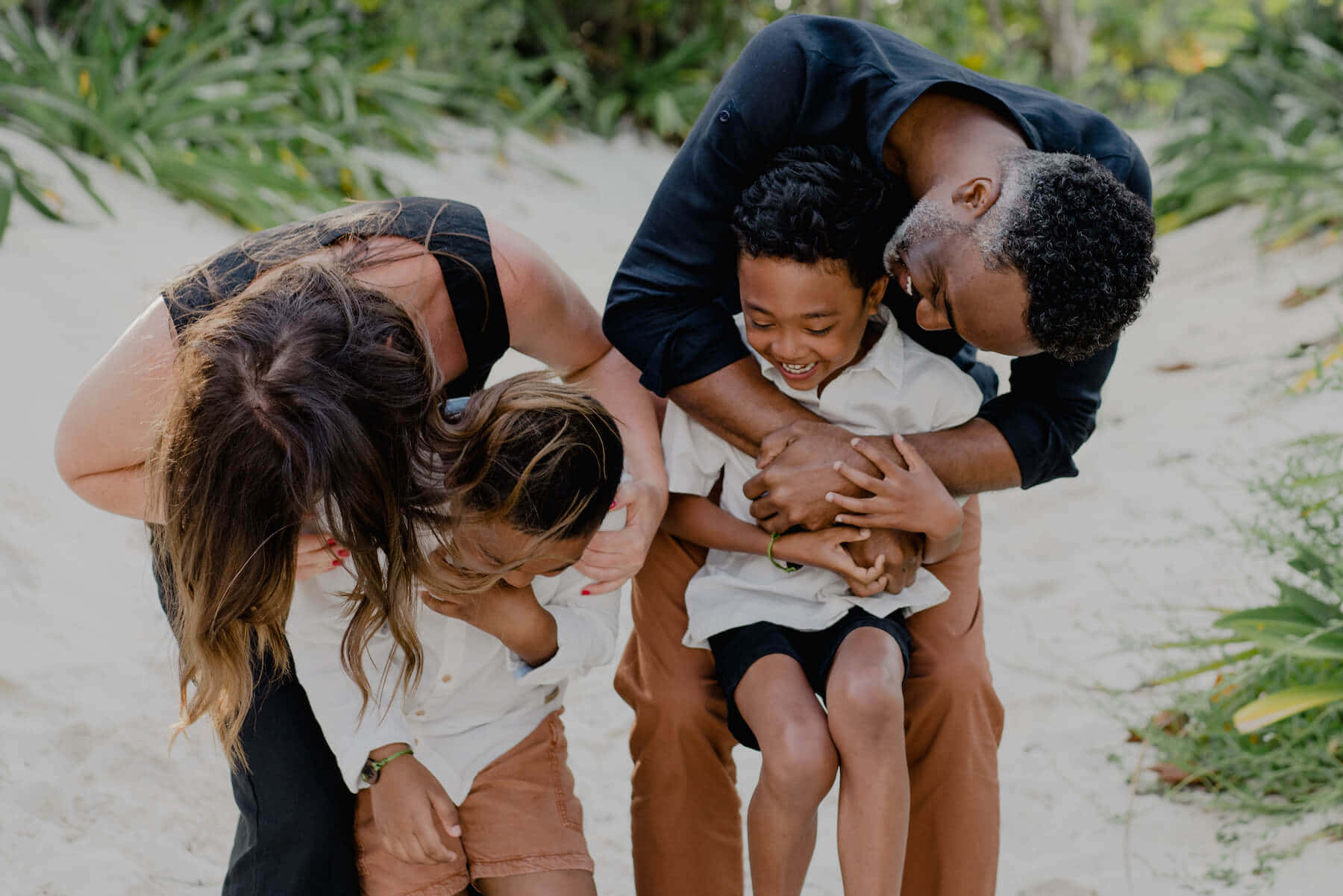 Family 桌布 壁紙 — 一個快樂的四口之家在陽光明媚的沙灘上擁抱，周圍是茂密的綠植。