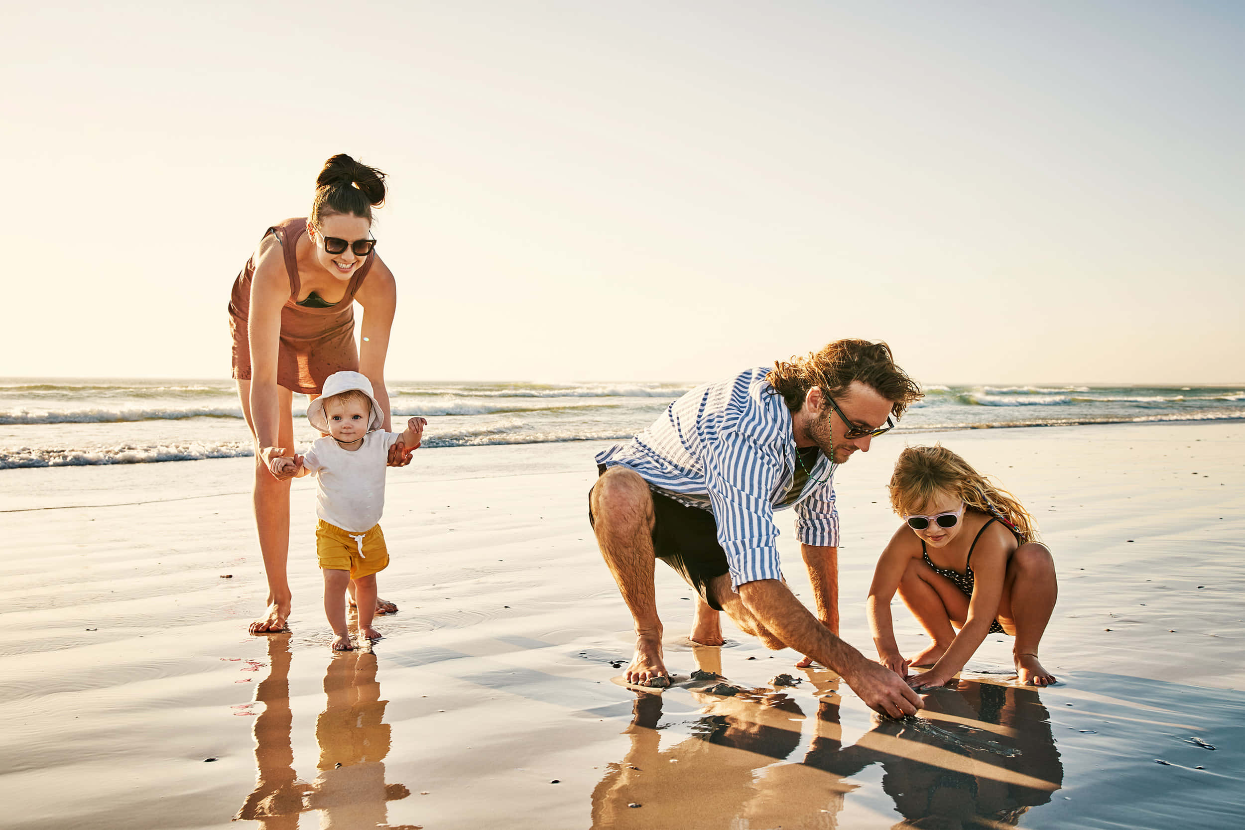 Beach Family 桌布 壁紙 — 一個快樂的四口之家在陽光明媚的海灘上一起玩耍，溫柔的海浪和濕沙反射出他們快樂的剪影。