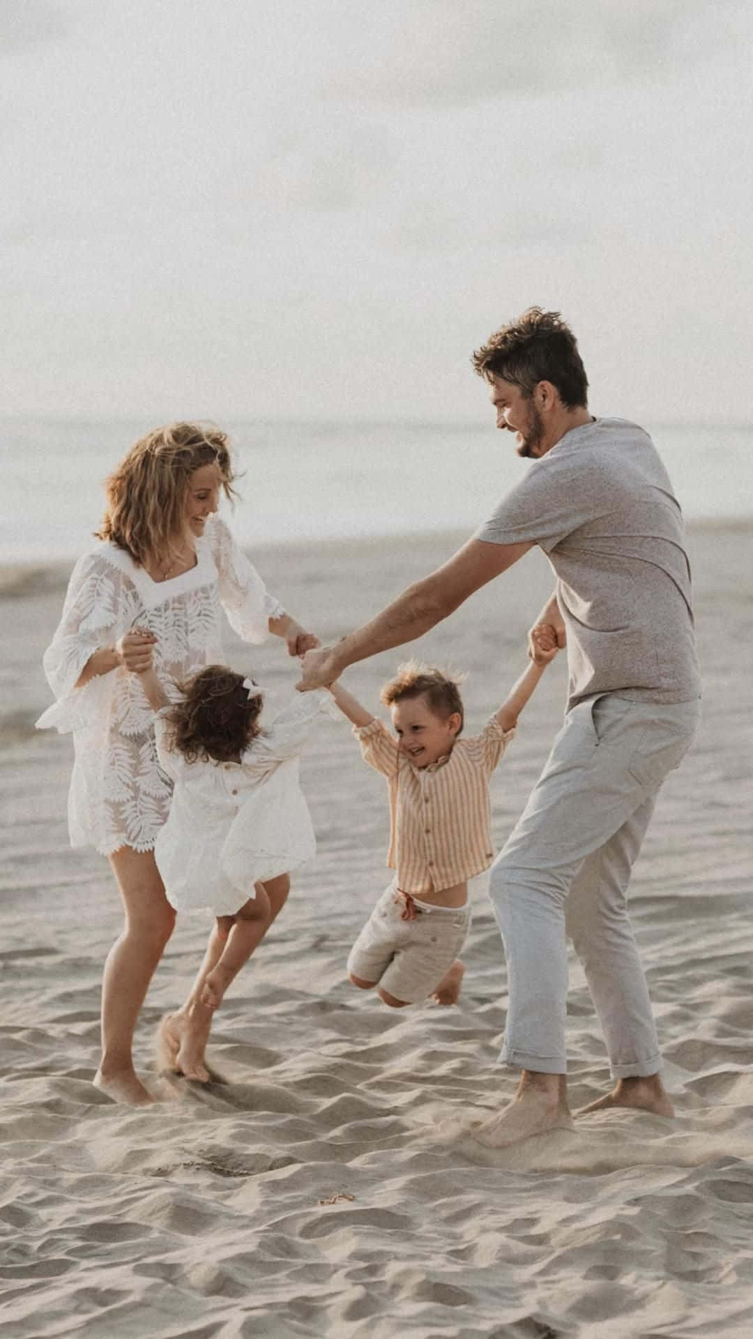 Family Beach 桌布 壁紙 — 一個快樂的四口之家在陽光明媚的海灘上旋轉，身後是柔軟的沙灘和寧靜的大海。