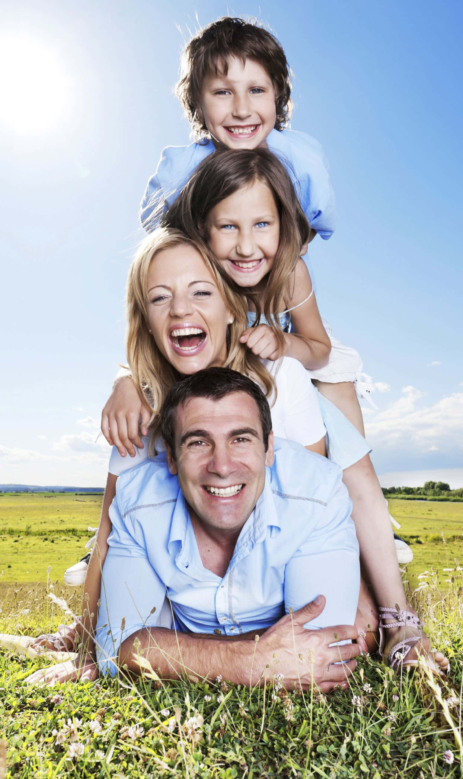 Family 桌布 壁紙 — 一個快樂的四口之家在陽光明媚的草地上，在明亮的藍天下一起大笑。