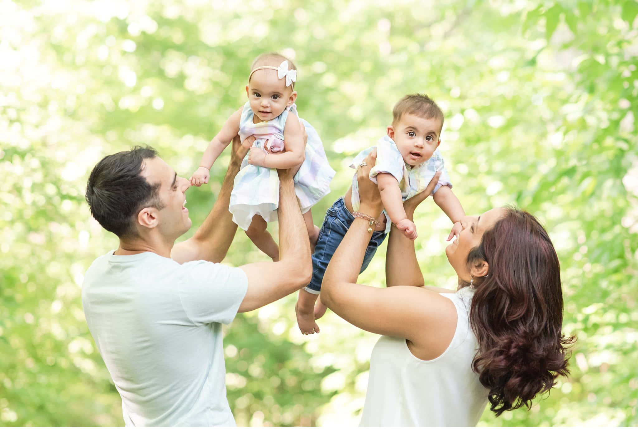 Spring Family 桌布 壁紙 — 一個快樂的四口之家在陽光普照、綠葉柔軟的花園中大笑，父母高舉他們的雙胞胎嬰兒。