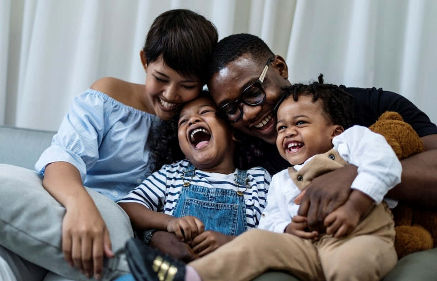 Black Family 桌布 壁紙 — 一個快樂的多種族四人家庭在沙發上一起大笑，身後是柔軟的白色窗簾。