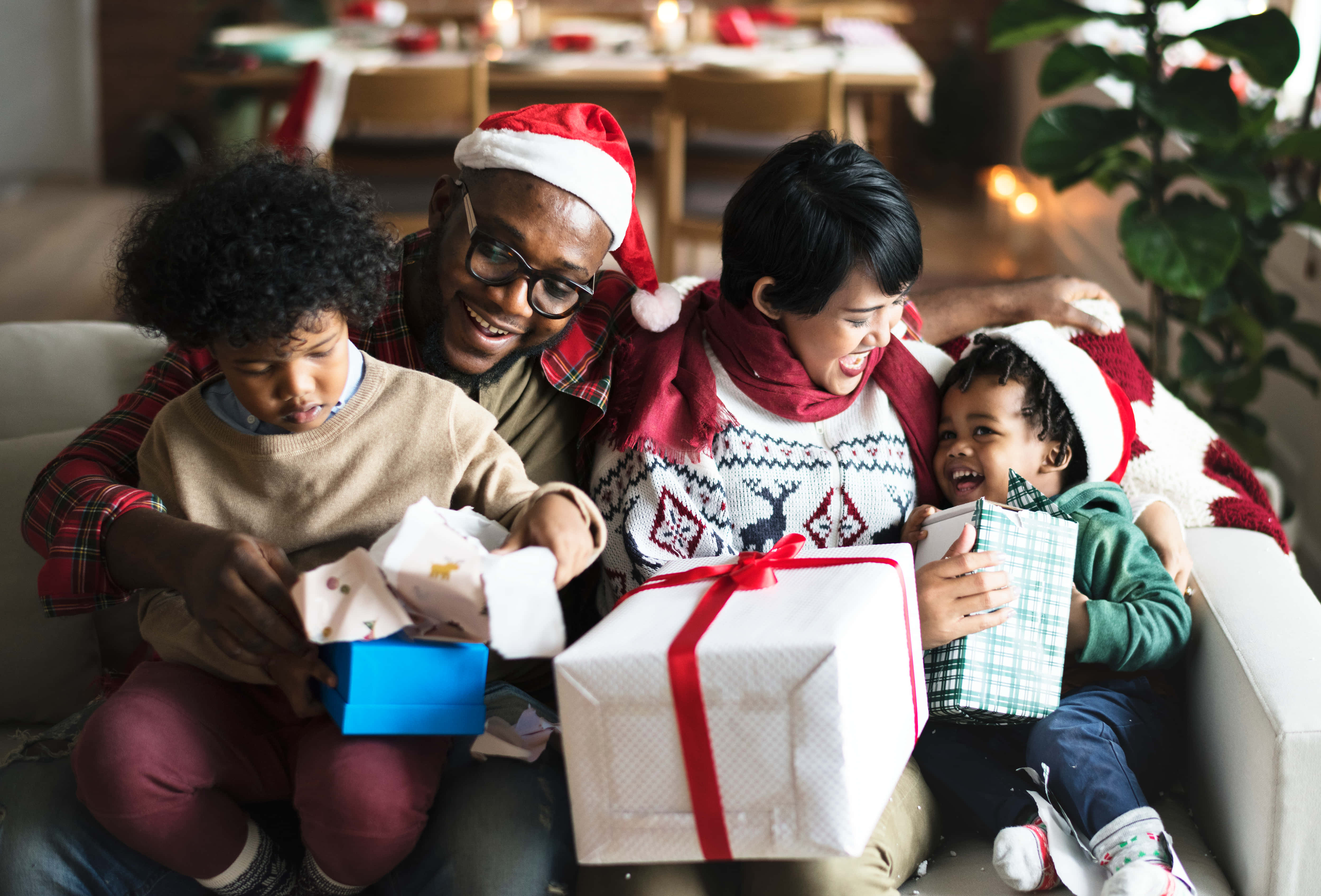 聖誕樹動畫 桌布 壁紙 — 一個快樂的多種族家庭穿著節日毛衣和聖誕帽，在舒適的沙發上一起打開聖誕禮物。