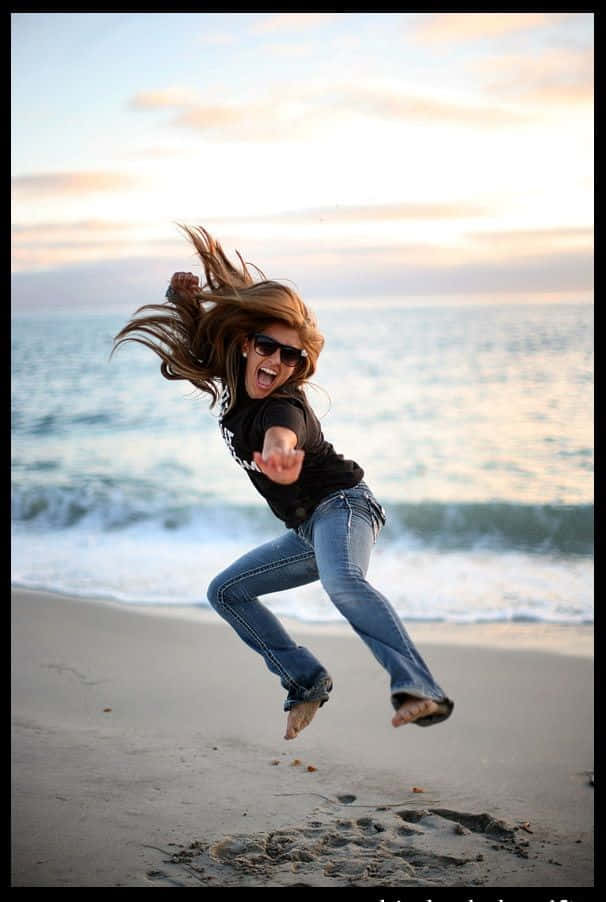 jump 桌布 壁紙 — 一個快樂的女人在日落時分赤腳跳躍在沙灘上，身後海浪拍打。